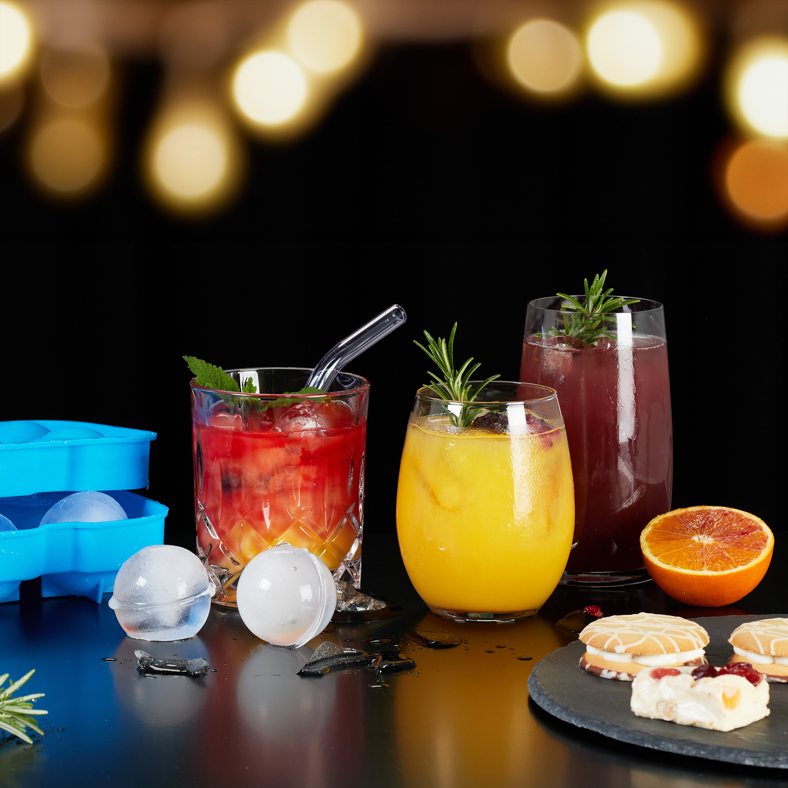 Drei bunte Cocktails mit Eiskugeln, Keksen und einer halben Orange auf dunkler Oberfläche vor Bokeh-Hintergrund.
