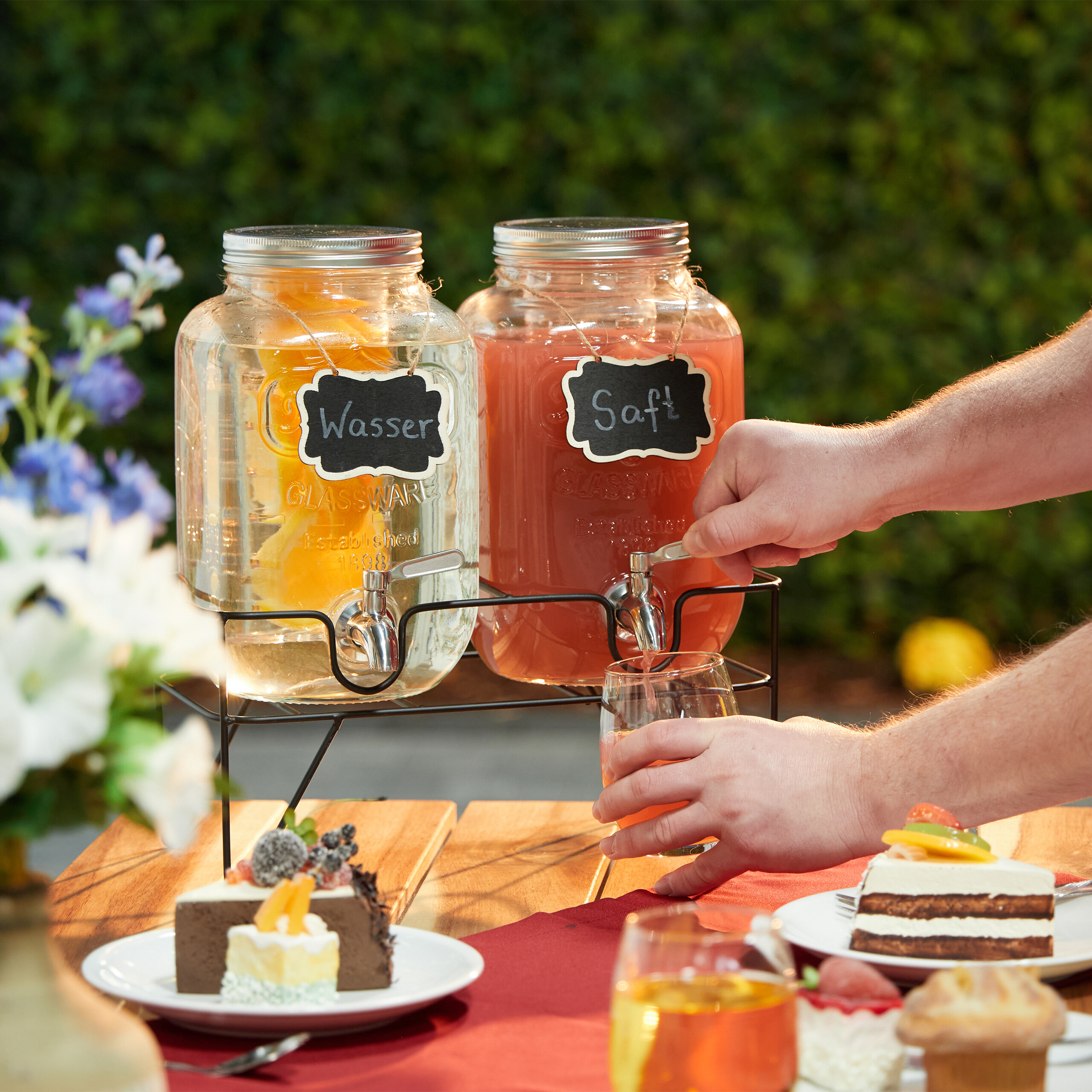 Person gießt Saft aus einem großen Glasbehälter auf einem Holztisch mit Kuchen und Blumen ein.
