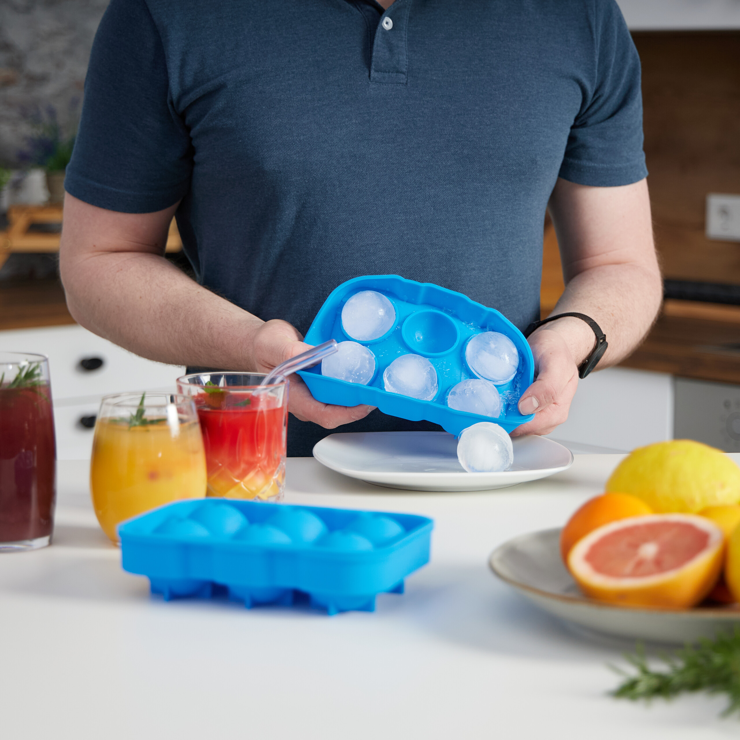 Ein Mann hält eine blaue Silikon-Eiswürfelform in der Küche, während Getränke und Früchte auf dem Tisch stehen.