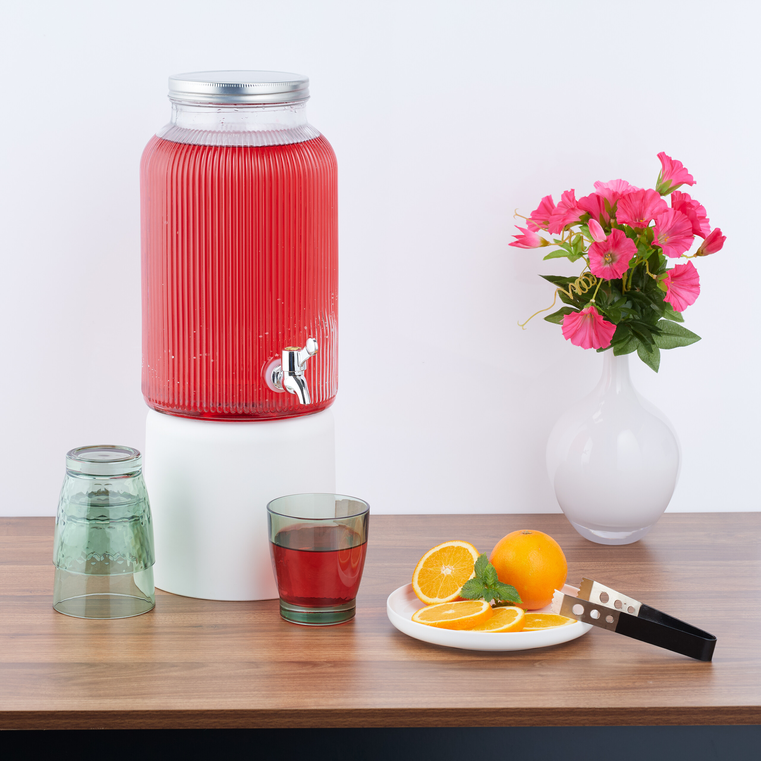 Ein roter Getränkespender, Gläser, Orangenscheiben auf einem Teller und rosa Blumen in einer weißen Vase auf einem Tisch.