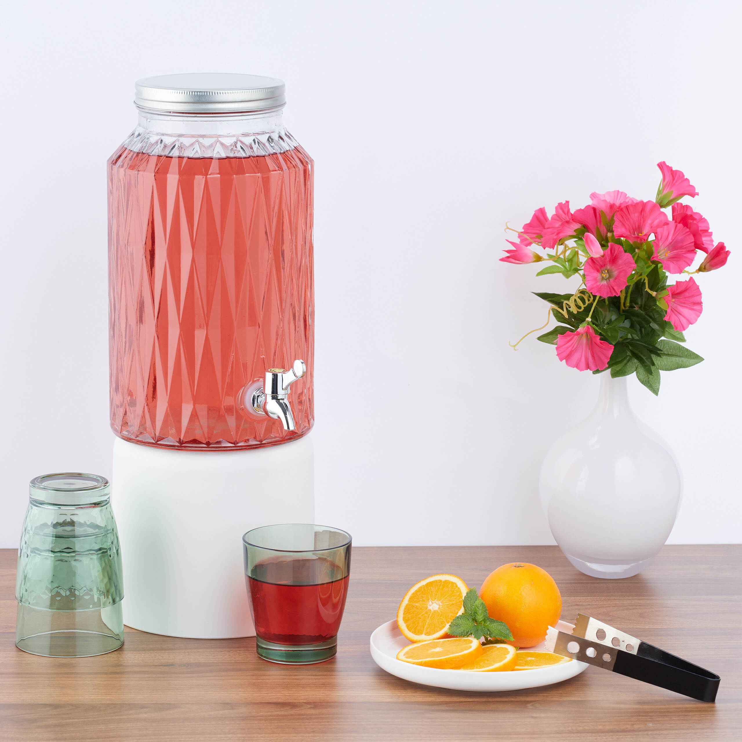 Ein großer roter Getränkespender, Gläser, Orangen auf einem Teller und rosa Blumen in einer Vase auf einem Holztisch.