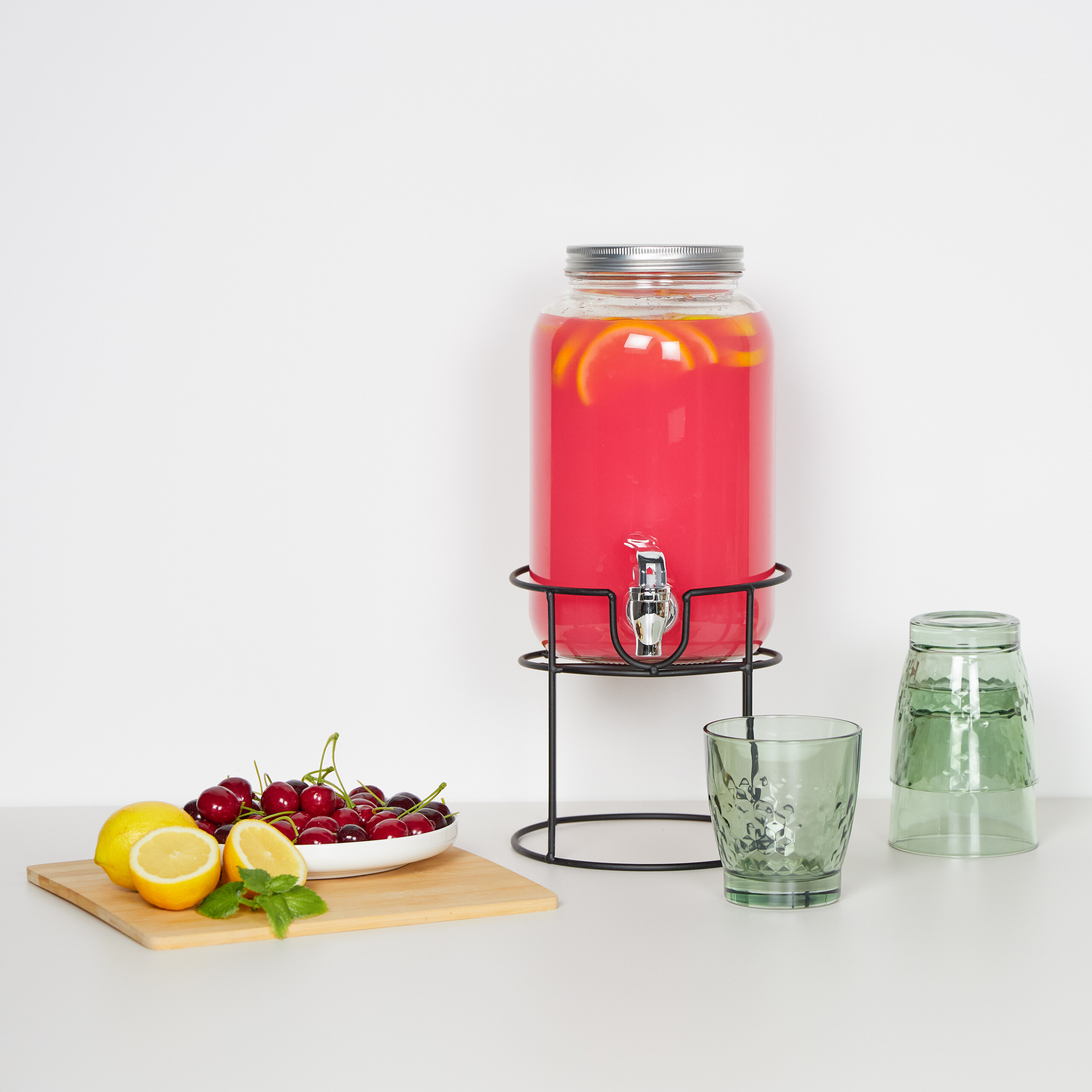 Ein Getränkespender mit pinker Limonade, Kirschen, Zitronen auf einem Holzbrett und grüne Gläser.