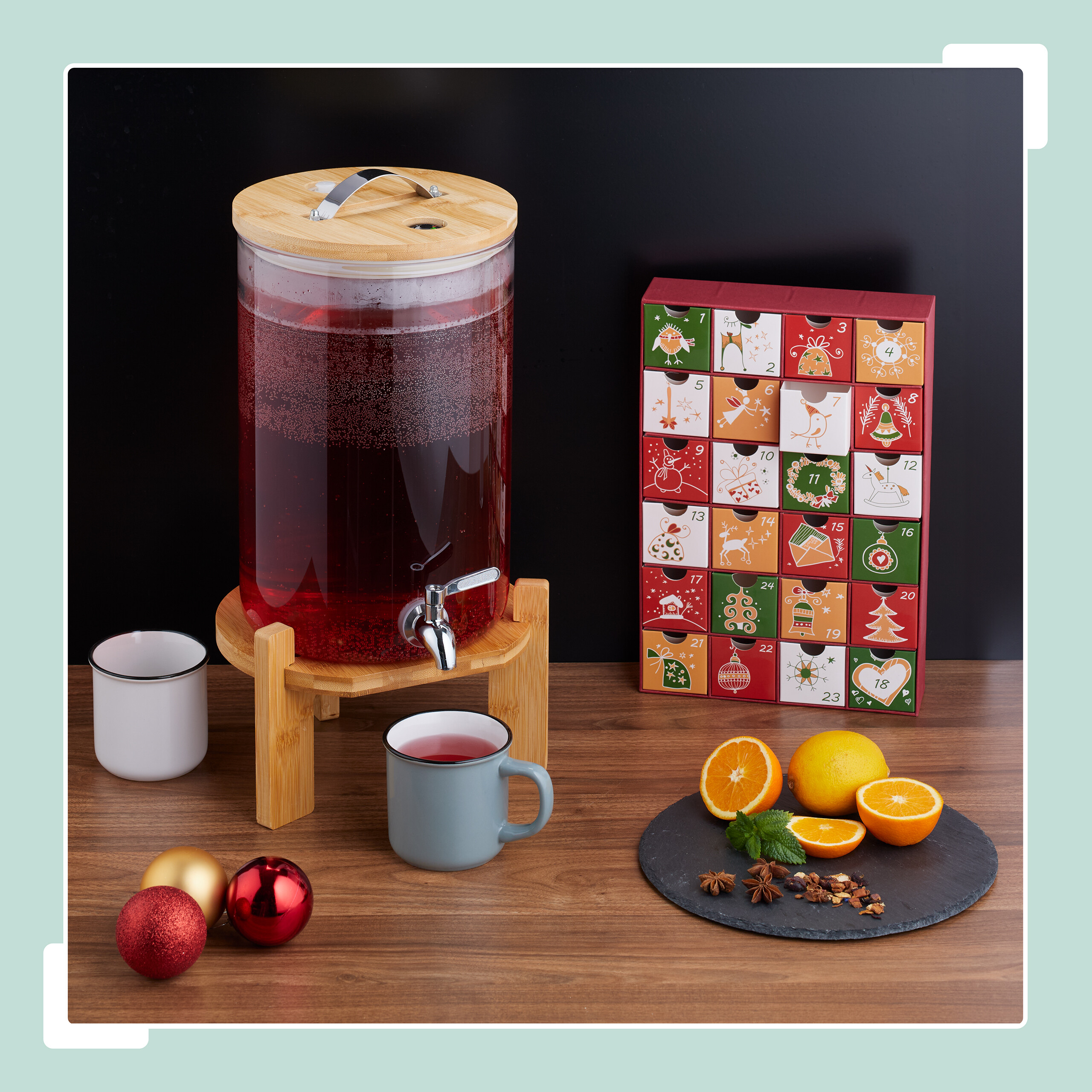 Ein Getränkespender mit rotem Saft steht auf Holztisch neben Adventskalender, Tassen und Orangenscheiben.