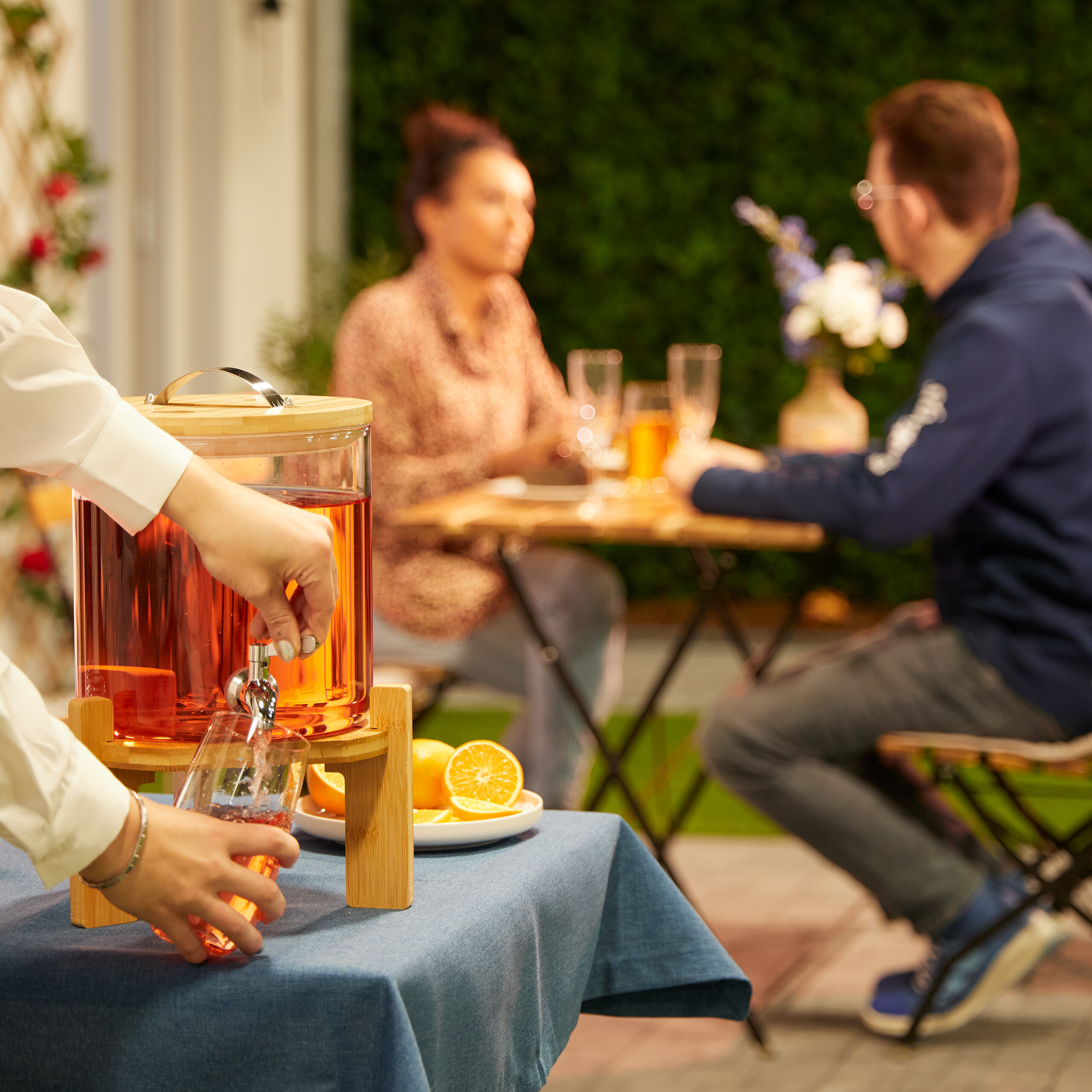Eine Person gießt Getränk aus einem Spender ein, während im Hintergrund ein Paar an einem Tisch sitzt.