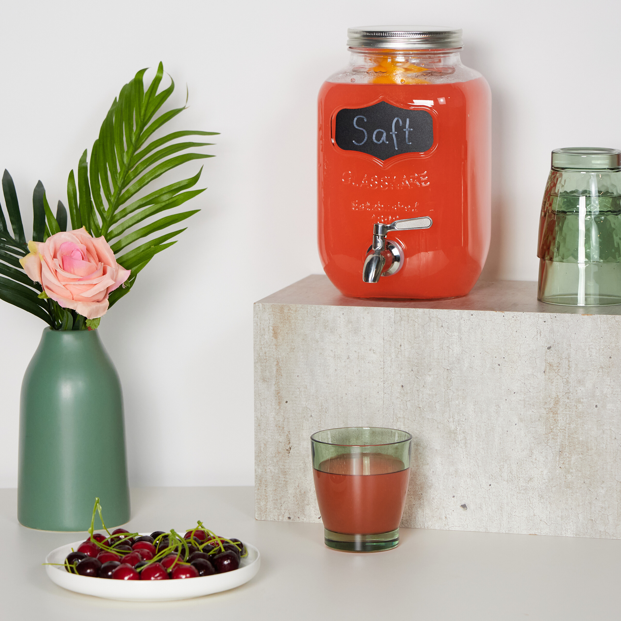 Ein roter Saftdispenser auf einem Sockel, daneben ein gefülltes Glas, Kirschen und eine Vase mit Blumen.