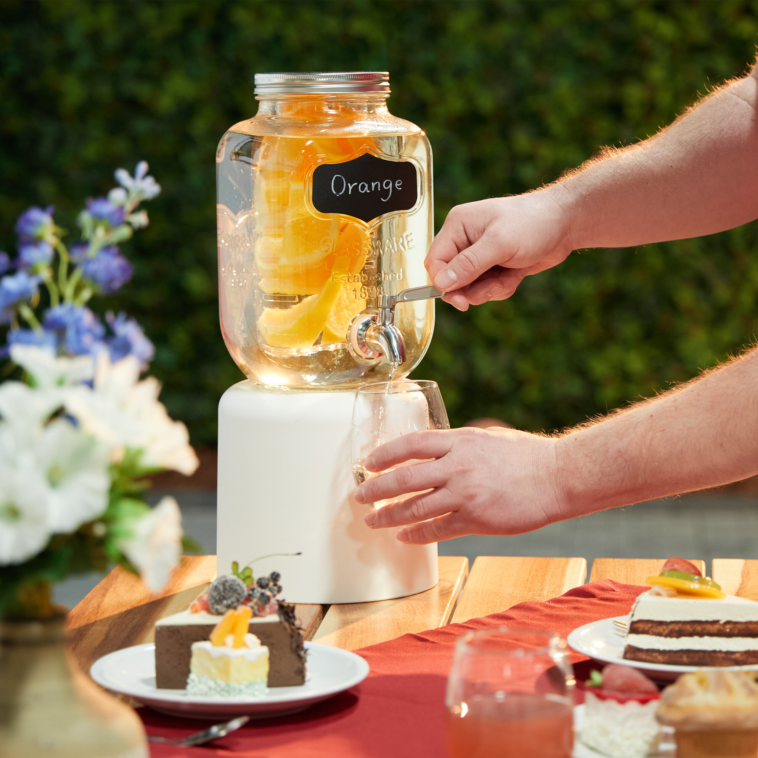 Jemand gießt Orangenwasser aus einem Glasdispenser in ein Glas, mit Desserts auf einem Tisch im Freien.