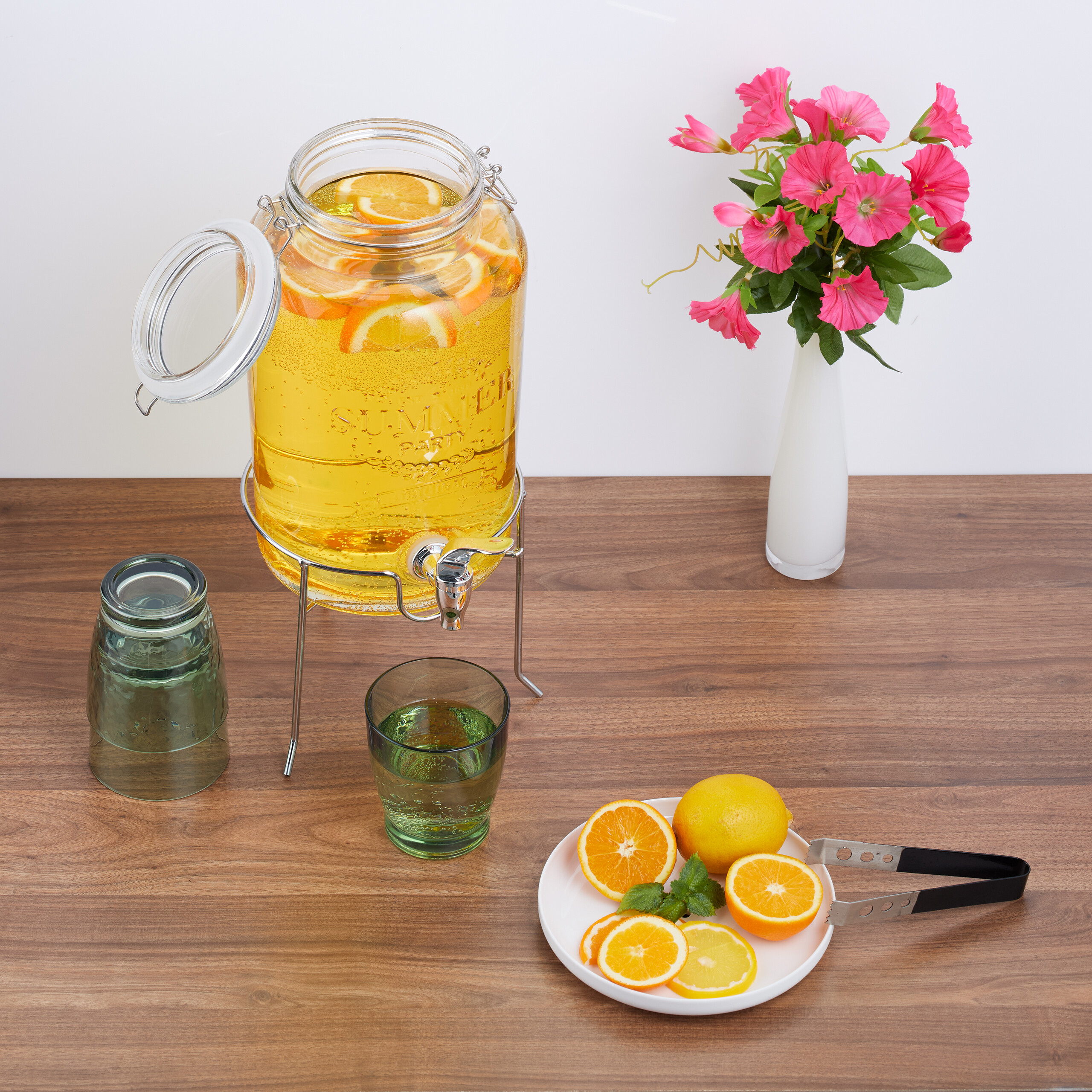 Ein Getränkespender mit Orangenscheiben steht neben Zitrusfrüchten, Gläsern und einer Blumenvase auf einem Holztisch.