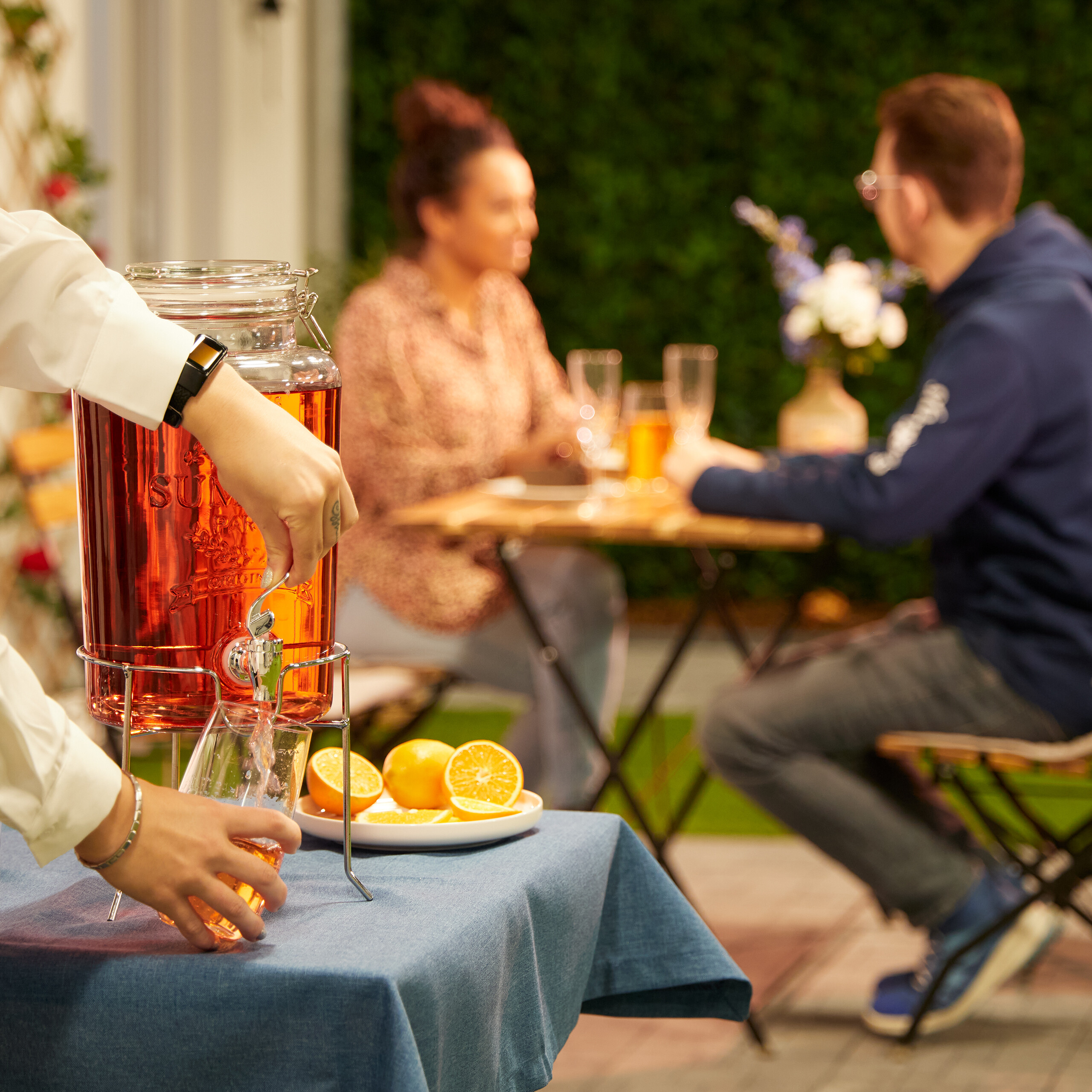 Eine Person gießt ein rosafarbenes Getränk aus einem Glasbehälter in ein Glas neben Orangenscheiben, mit einem Paar im Hintergrund.