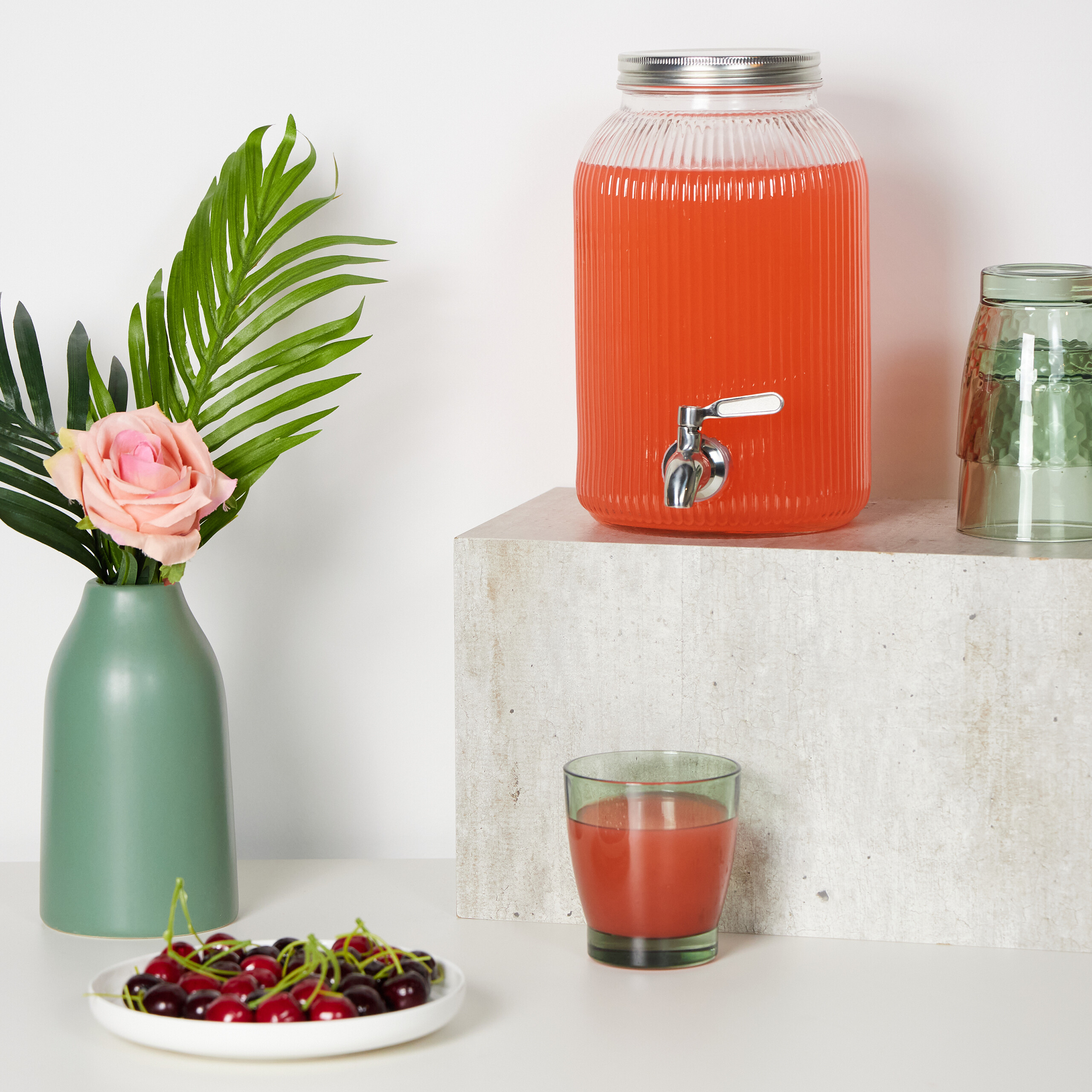 Ein großer Glasdispenser mit rosa Saft steht auf einem Steinblock, neben einer Vase mit rosa Rose, Kirschen und Gläsern.