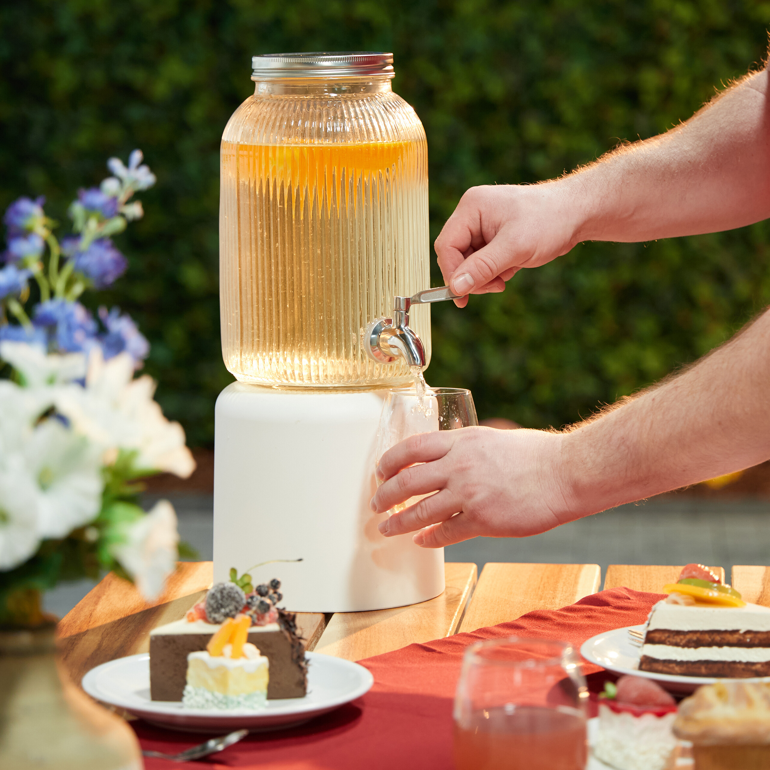 Eine Person zapft Getränk aus einem großen Glastopf im Garten, umgeben von Kuchen.