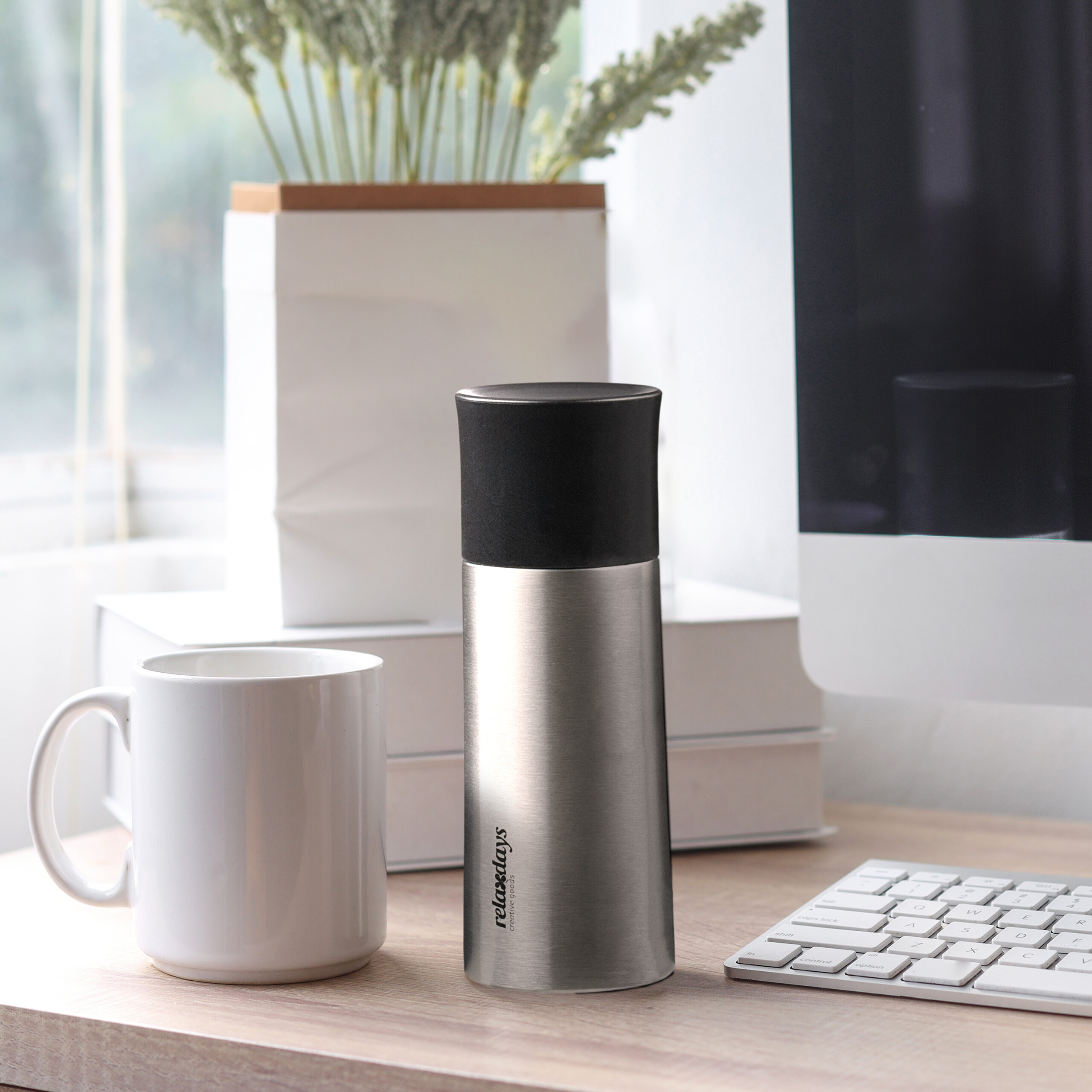 Edelstahl-Thermoskanne auf einem Schreibtisch neben einer Kaffeetasse, Büchern und einem Computer.