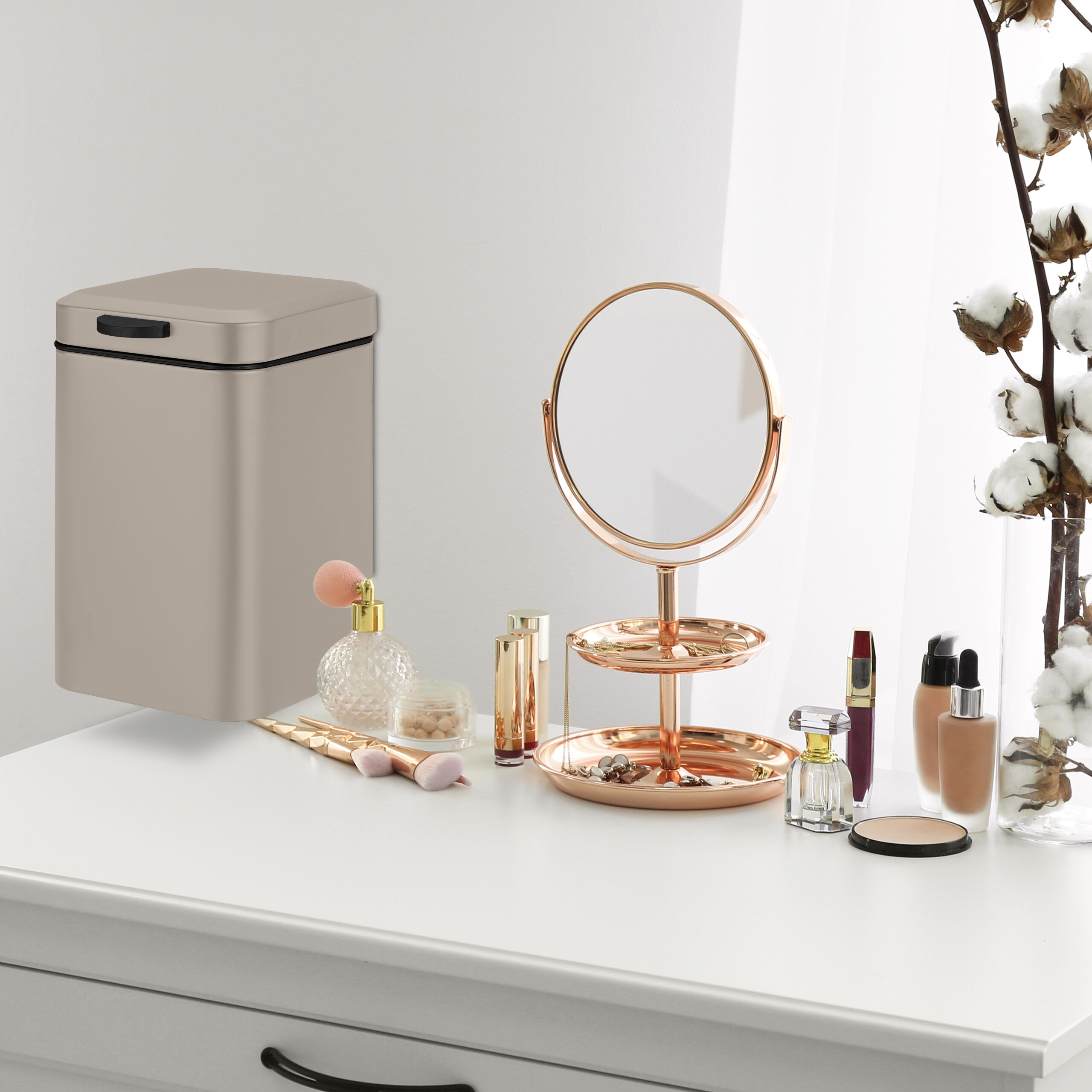 Ein Kosmetiktisch mit Rosegold-Spiegel, Schmuck, Make-up und einem kleinen beigen Mülleimer.