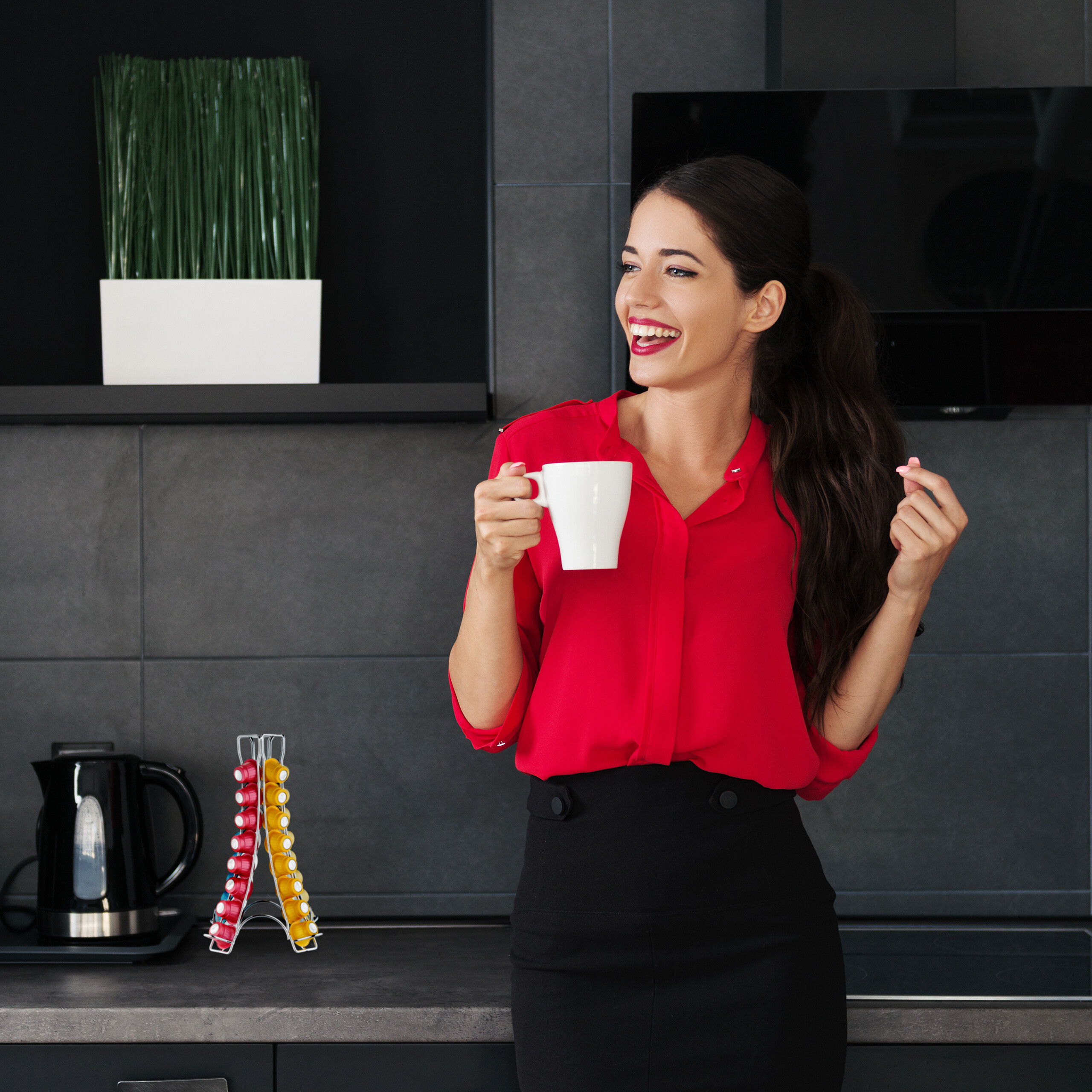 Eine lächelnde Frau in rotem Hemd und schwarzem Rock hält eine weiße Tasse in einer modernen Küche.