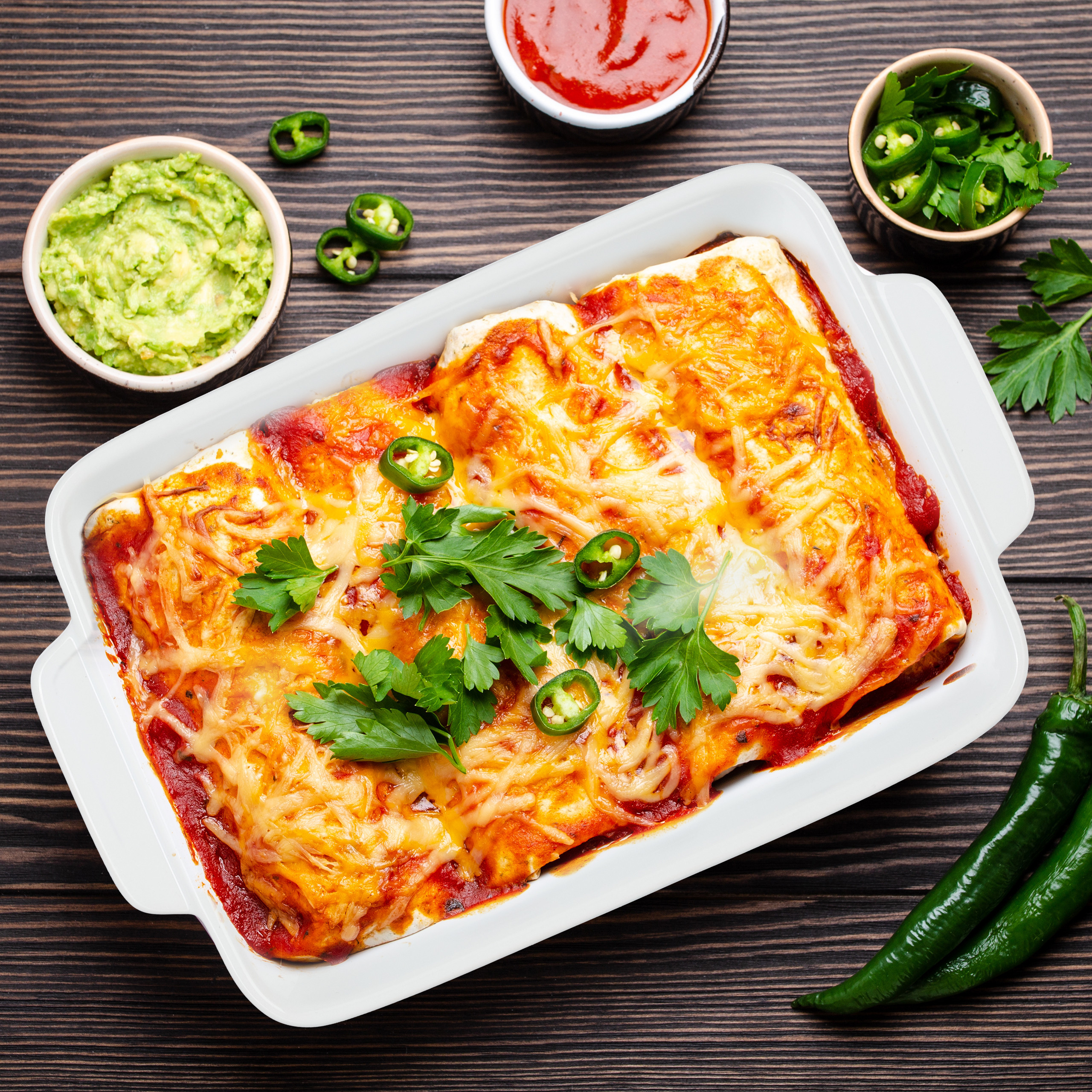 Gebackene Enchiladas in einer weißen Schale mit Käse, Tomatensoße, Petersilie und Jalapeños, mit Guacamole und Salsa auf Holz.