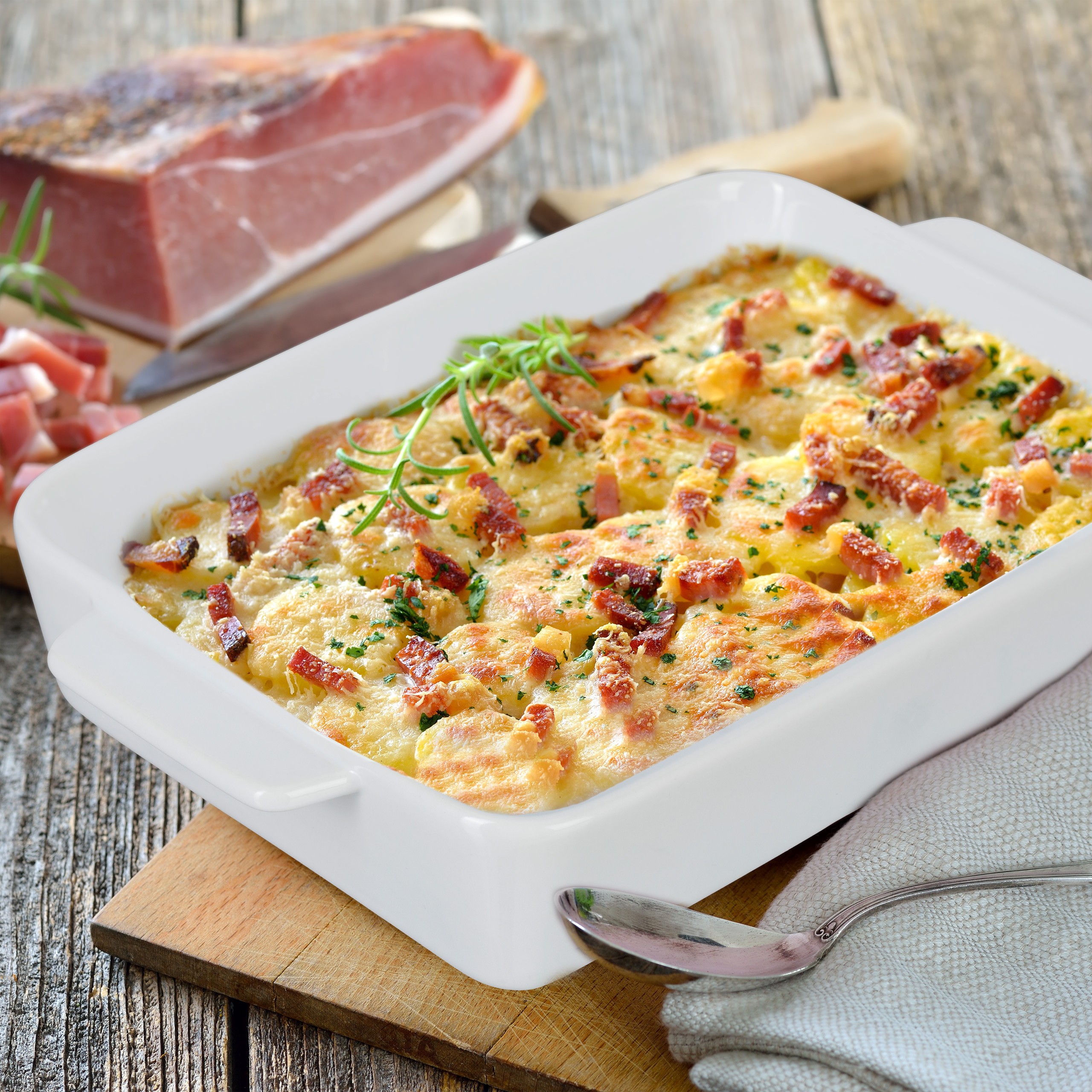Goldbrauner Kartoffelauflauf mit Speck in weißer Auflaufform auf Holzbrett, im Hintergrund Schinken.