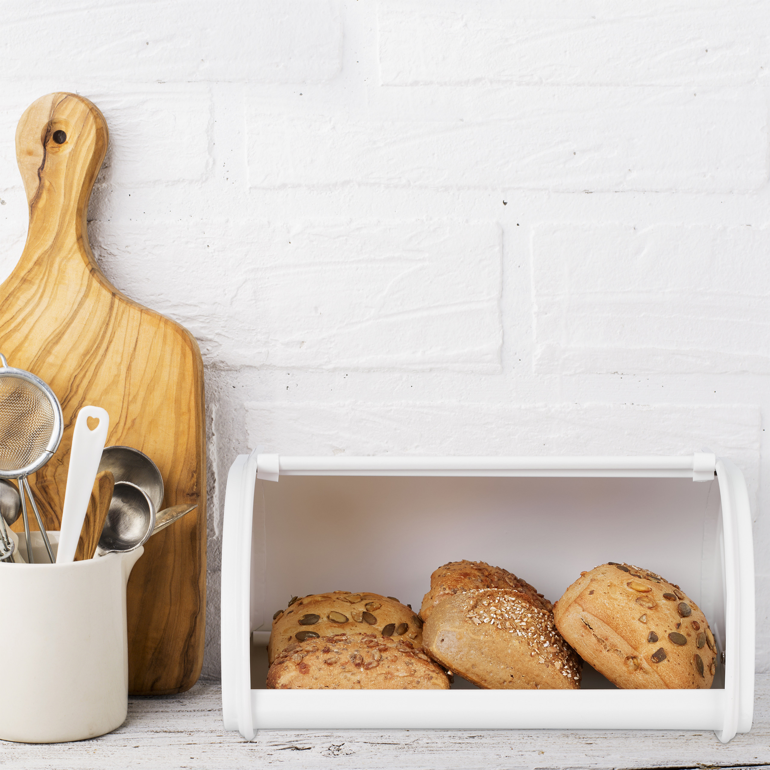 Verschiedene Brötchen in einem weißen Brotkasten, daneben ein Holzschneidebrett und Utensilien.