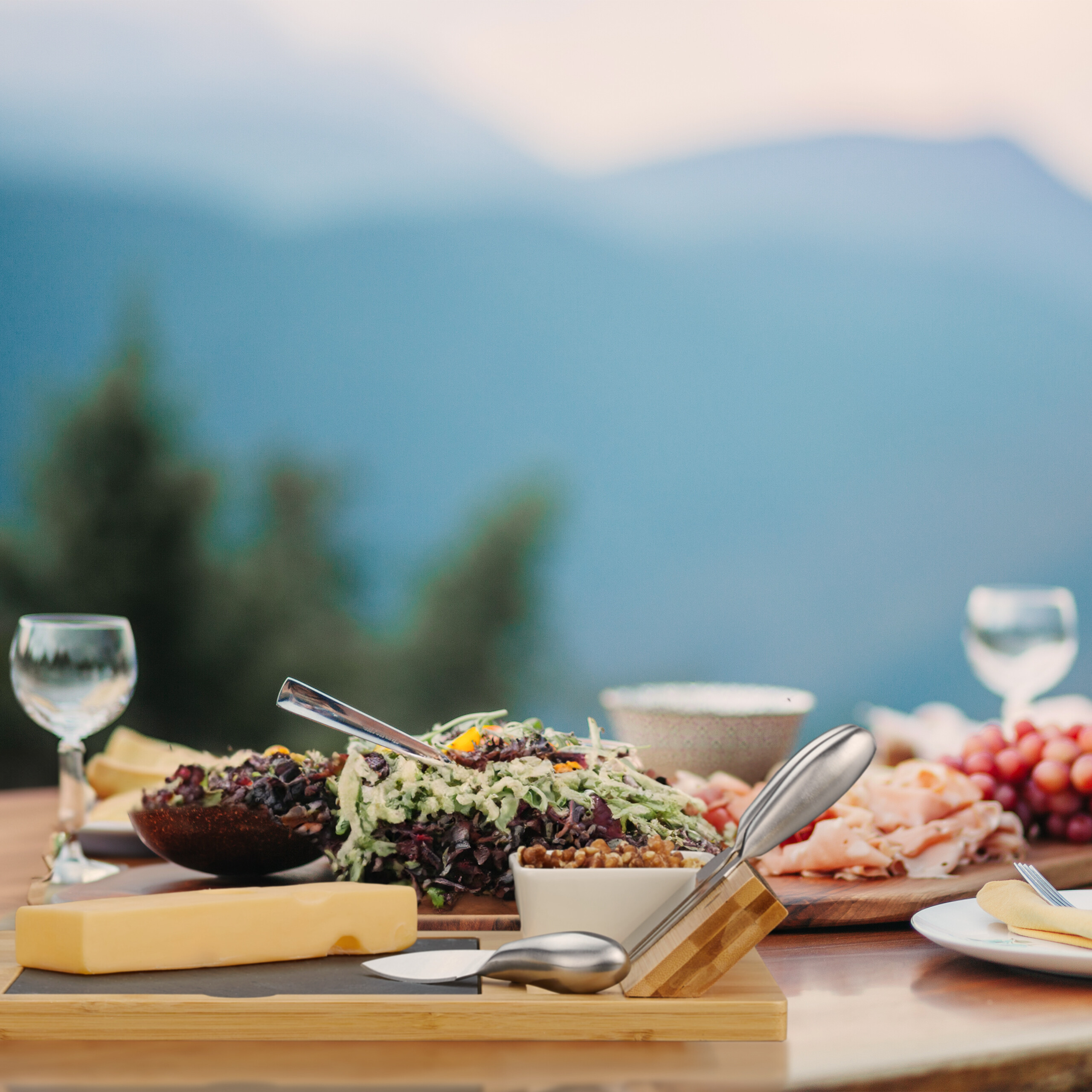 Ein Tisch mit Salat, Käse und Weingläsern vor einer Bergkulisse.