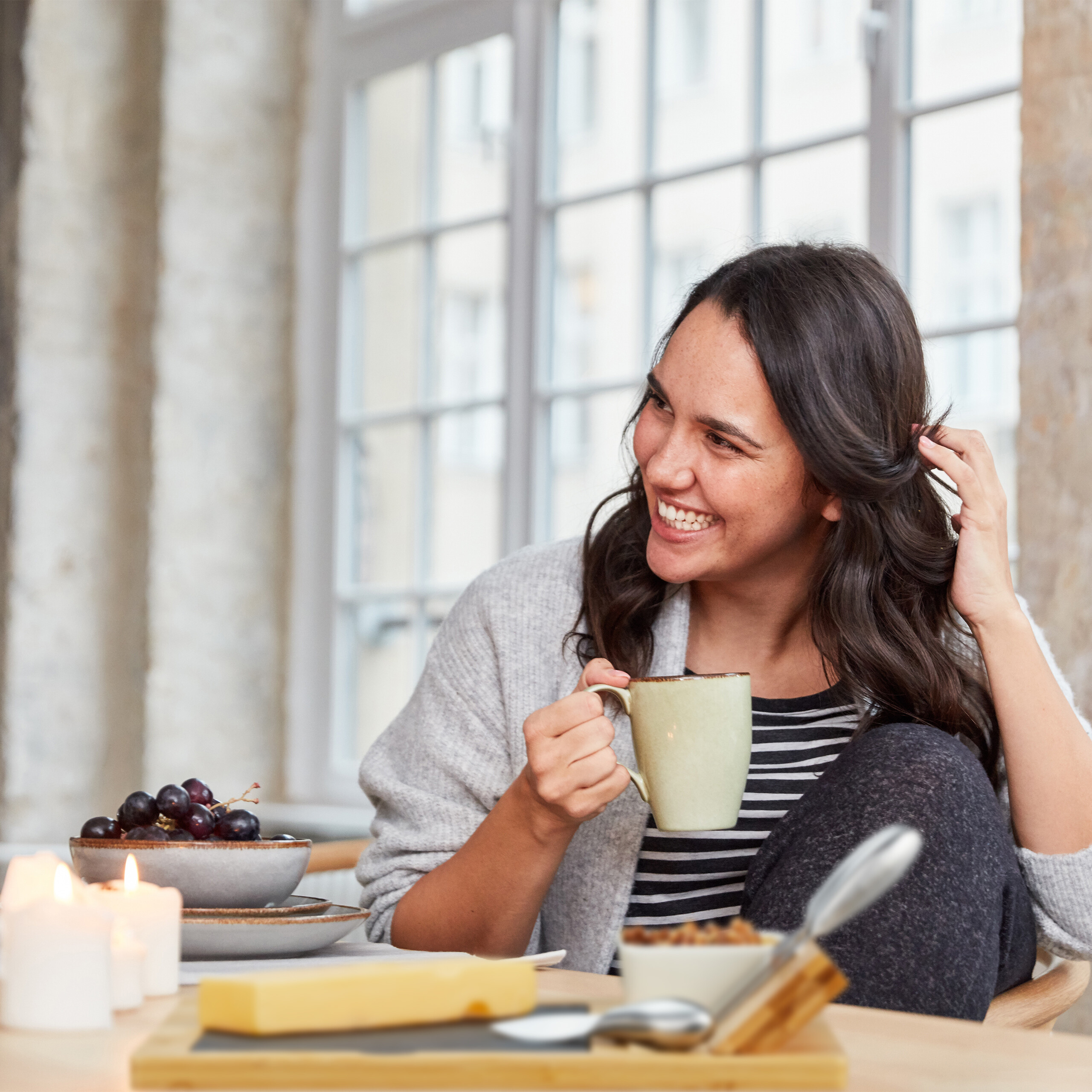 Eine lächelnde Frau hält eine Tasse, an einem Tisch mit Trauben, Käse und Kerzen vor einem hellen Fenster.