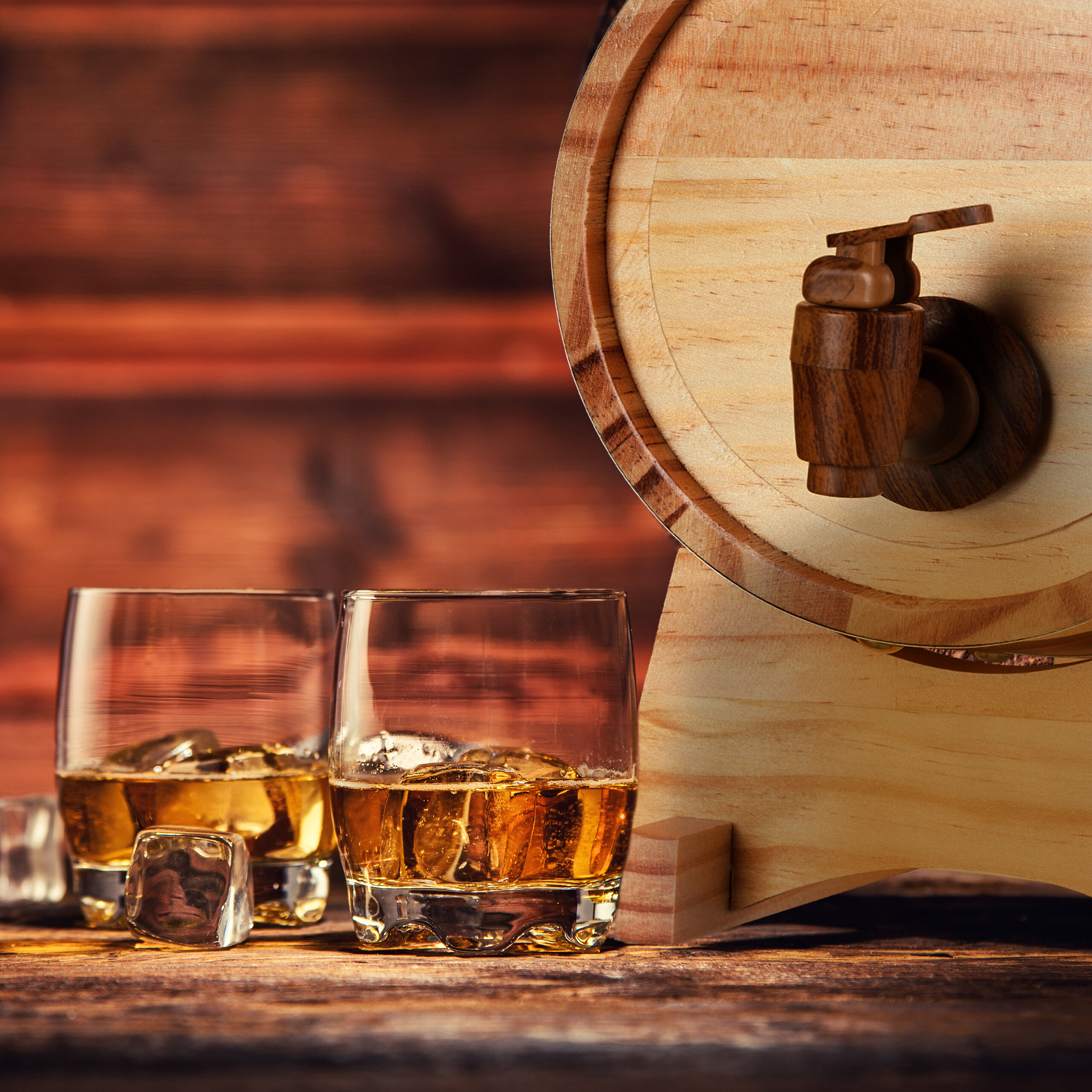 Zwei Gläser Whiskey mit Eiswürfeln stehen vor einem kleinen Holzfass auf einer Holztischplatte.