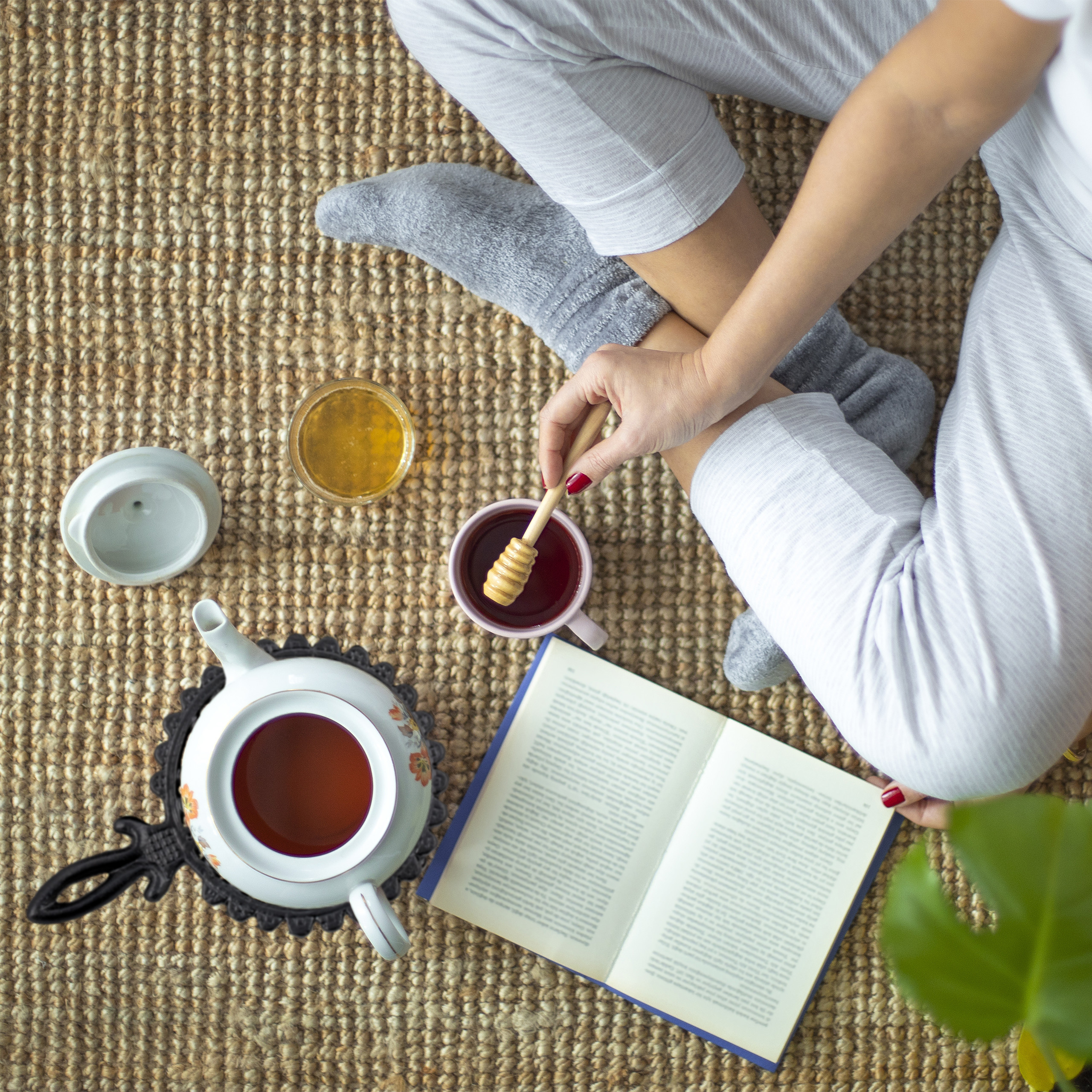 Person in grauen Socken sitzt auf Teppich mit Teekanne, Tasse Honig, offenem Buch und Teetasse mit Honiglöffel.