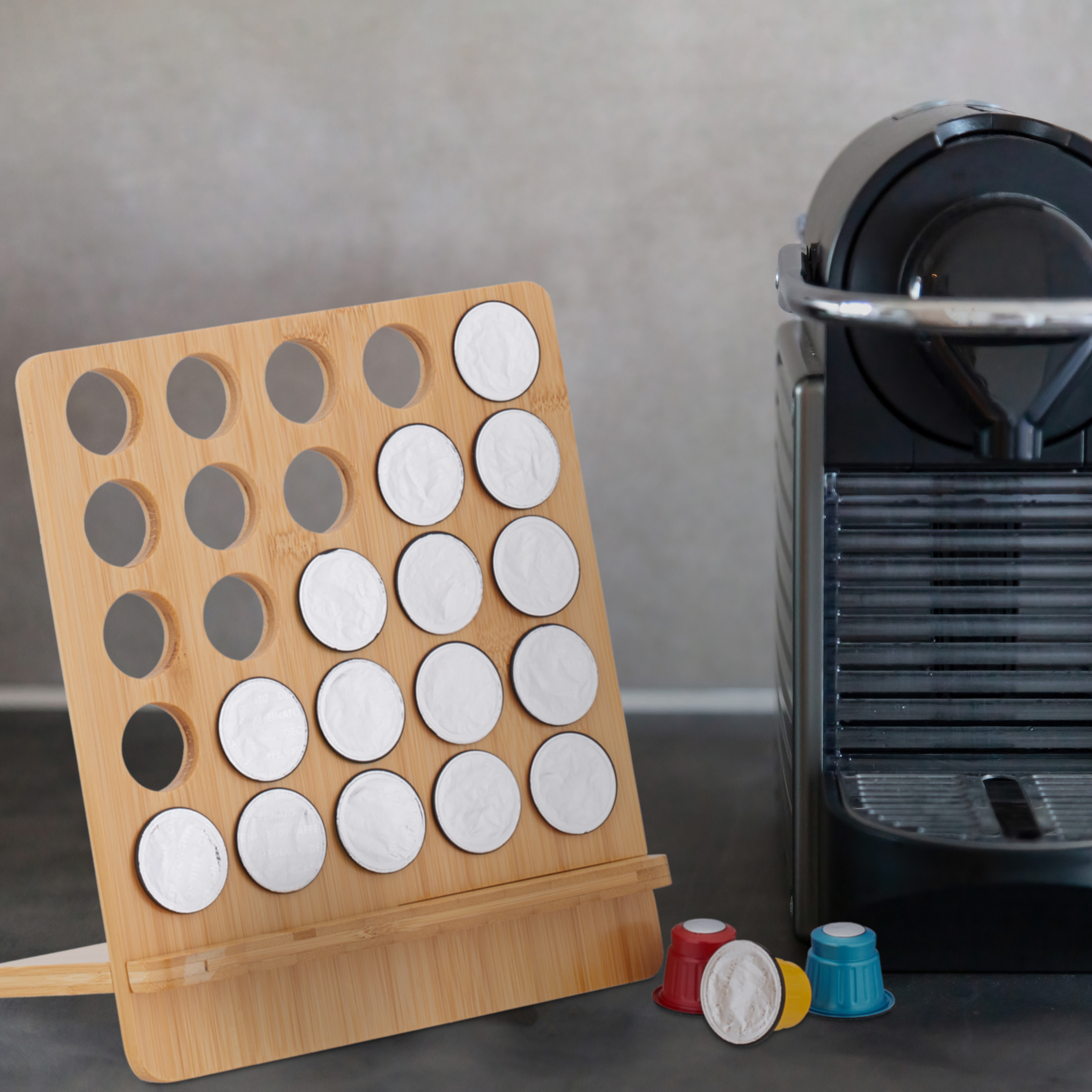 Ein hölzerner Kaffeekapselhalter mit Kapseln steht vor einer grauen Kaffeemaschine auf dunkler Oberfläche.