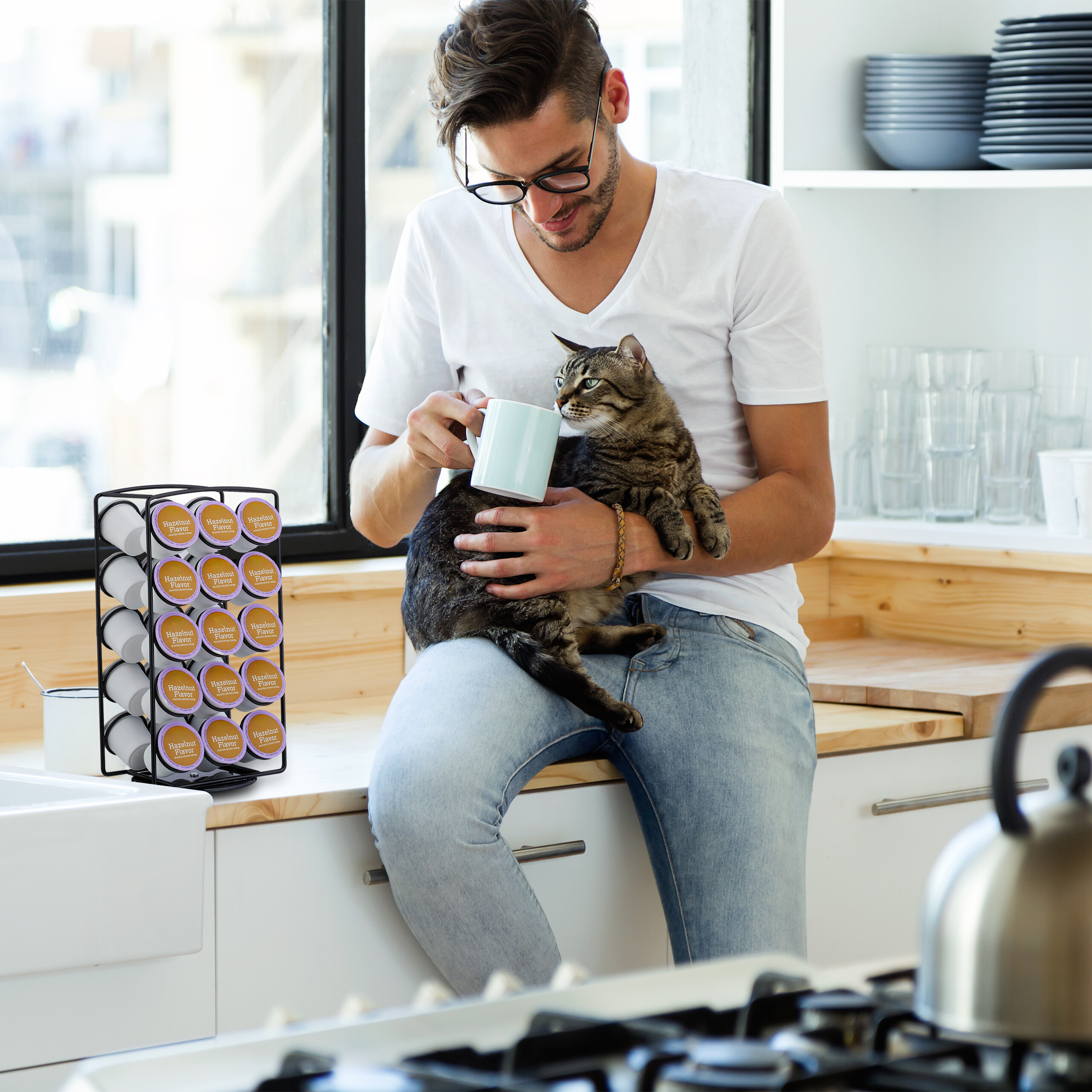 Ein Mann im weißen Hemd sitzt in einer Küche, hält eine Katze und reicht ihr eine Tasse.