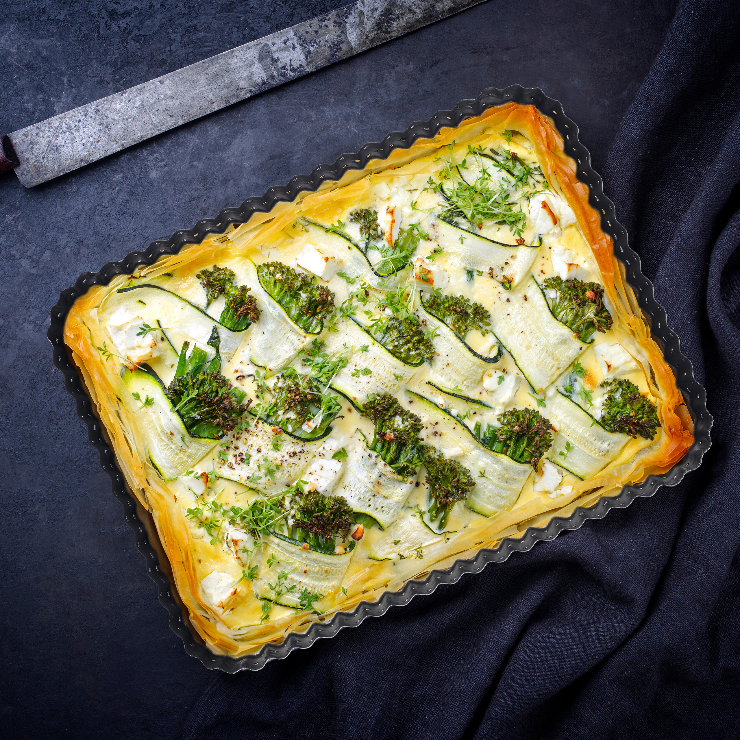 Eine frische Gemüse-Tarte mit Zucchini, Brokkoli und Feta in einer Form auf dunklem Untergrund.