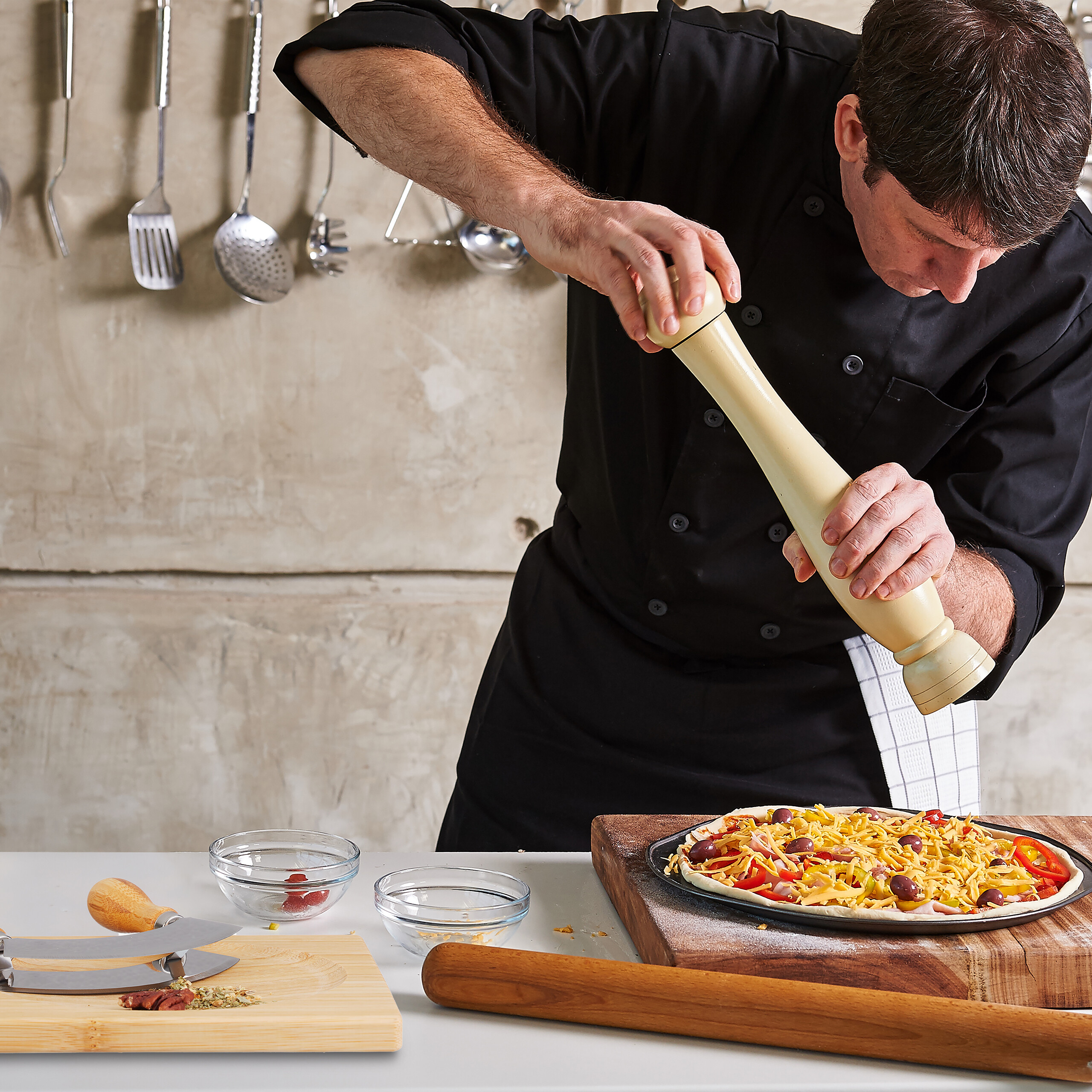 Ein Koch im schwarzen Uniform mahlt Gewürze auf eine rohe Pizza mit Käse und Oliven auf einem Holztisch in der Küche.