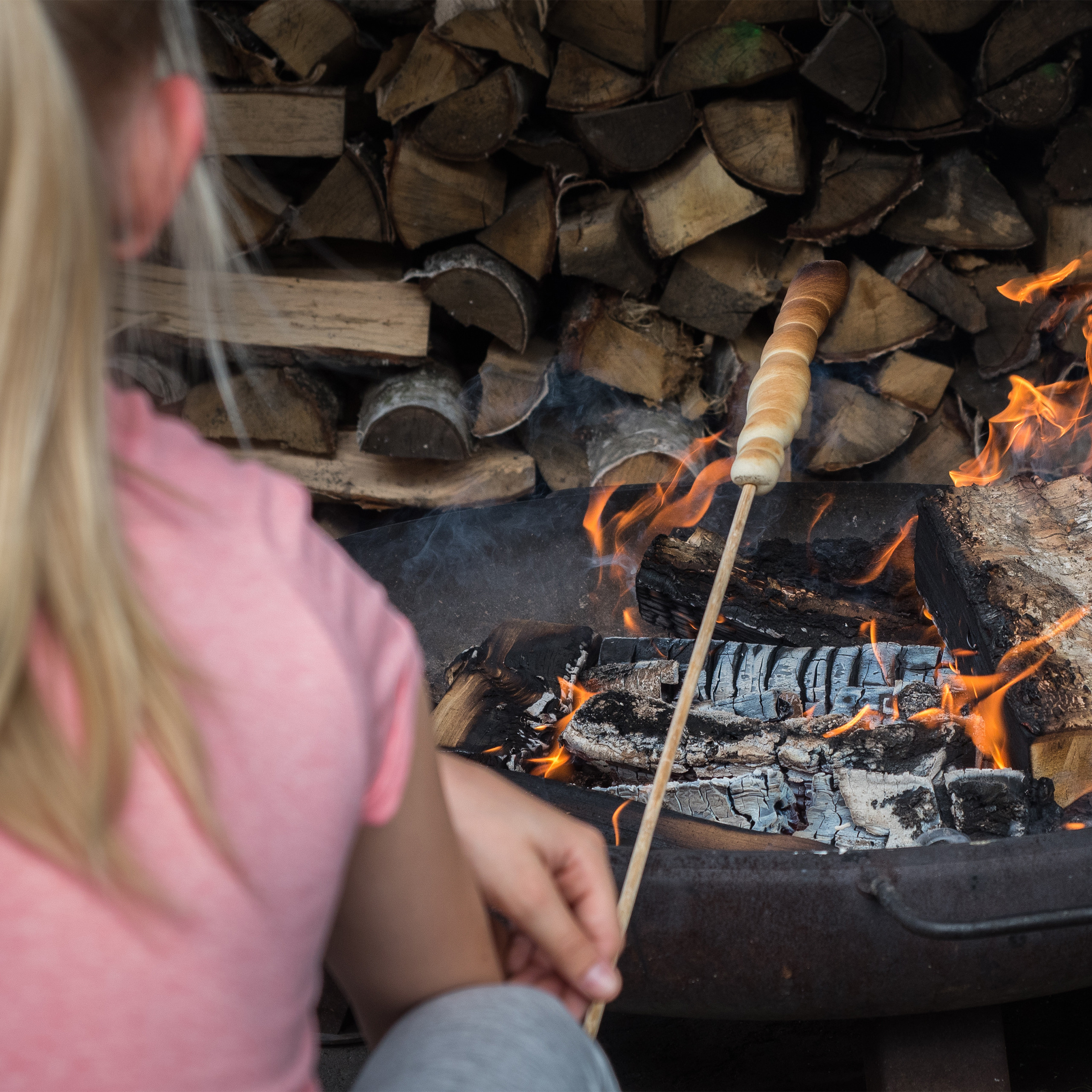 Ein blondes Mädchen röstet Brot an einem Stock über einer Feuerstelle mit Holzstapel im Hintergrund.