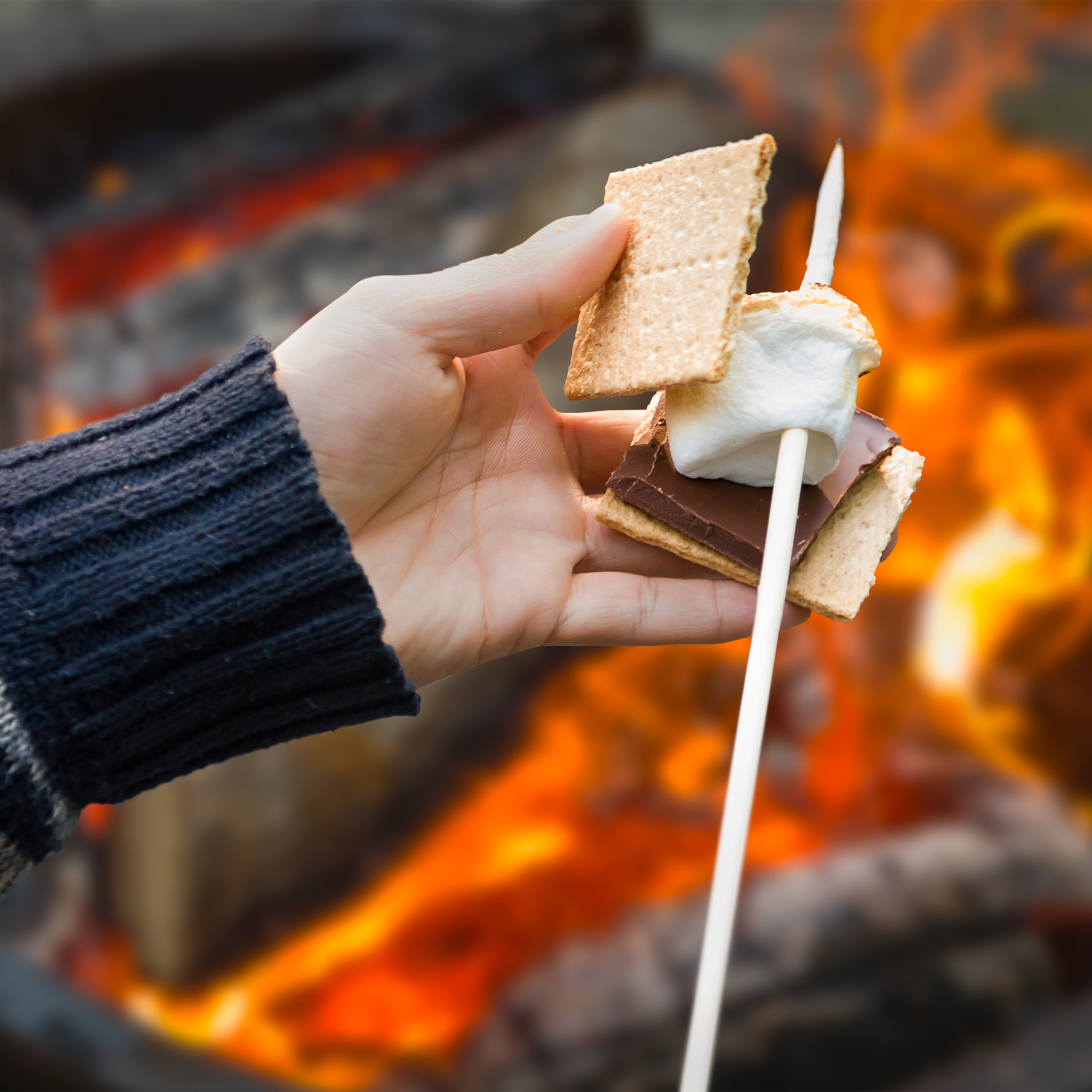 Eine Hand hält einen Marshmallow und Schokolade zwischen Crackern über einem Lagerfeuer.