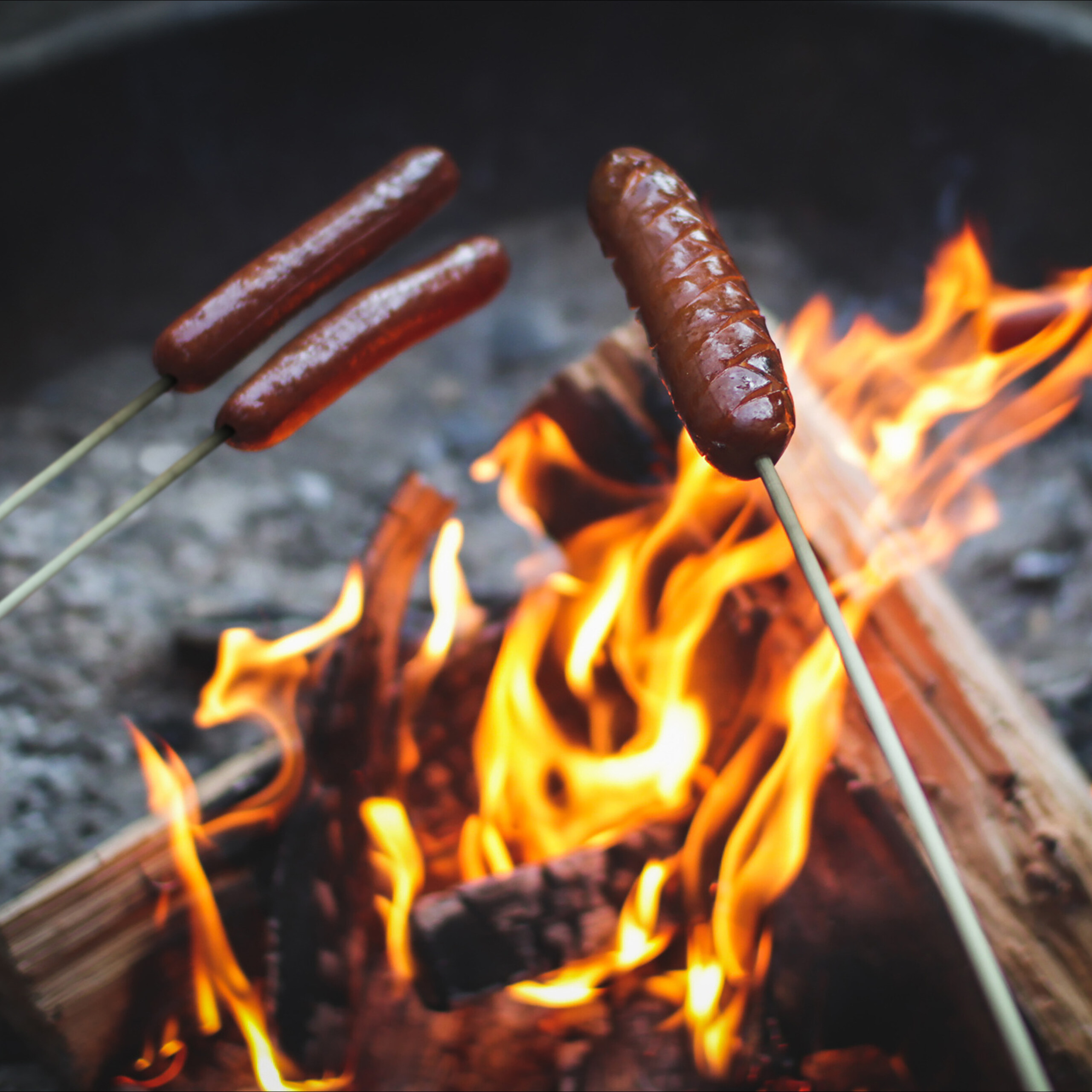 Drei Würstchen garen an Holzspießen über einem offenen Lagerfeuer.