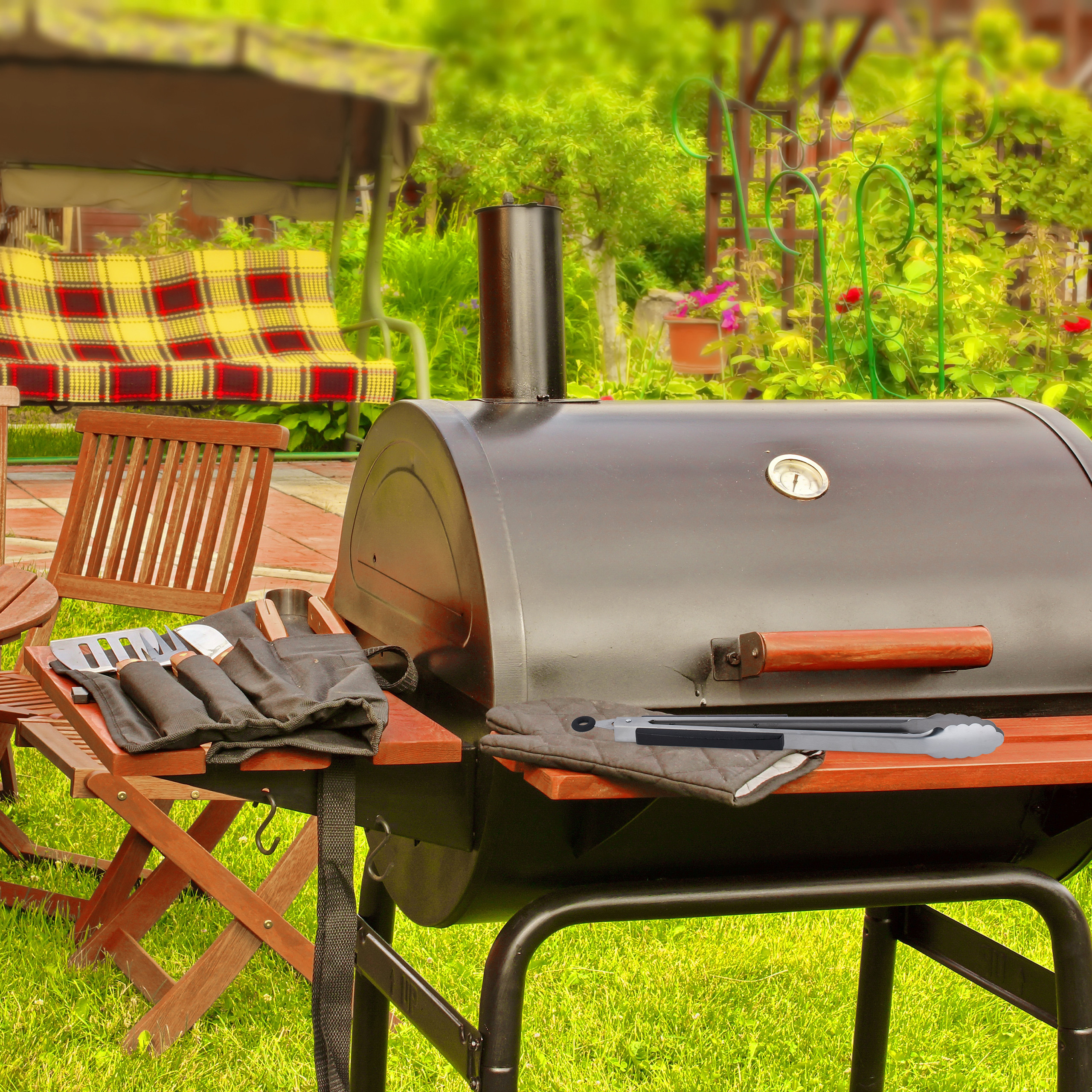 Ein schwarzer Grill mit Thermometer steht auf Gras, daneben Holztische und Stühle sowie ein farbiger Pavillon.