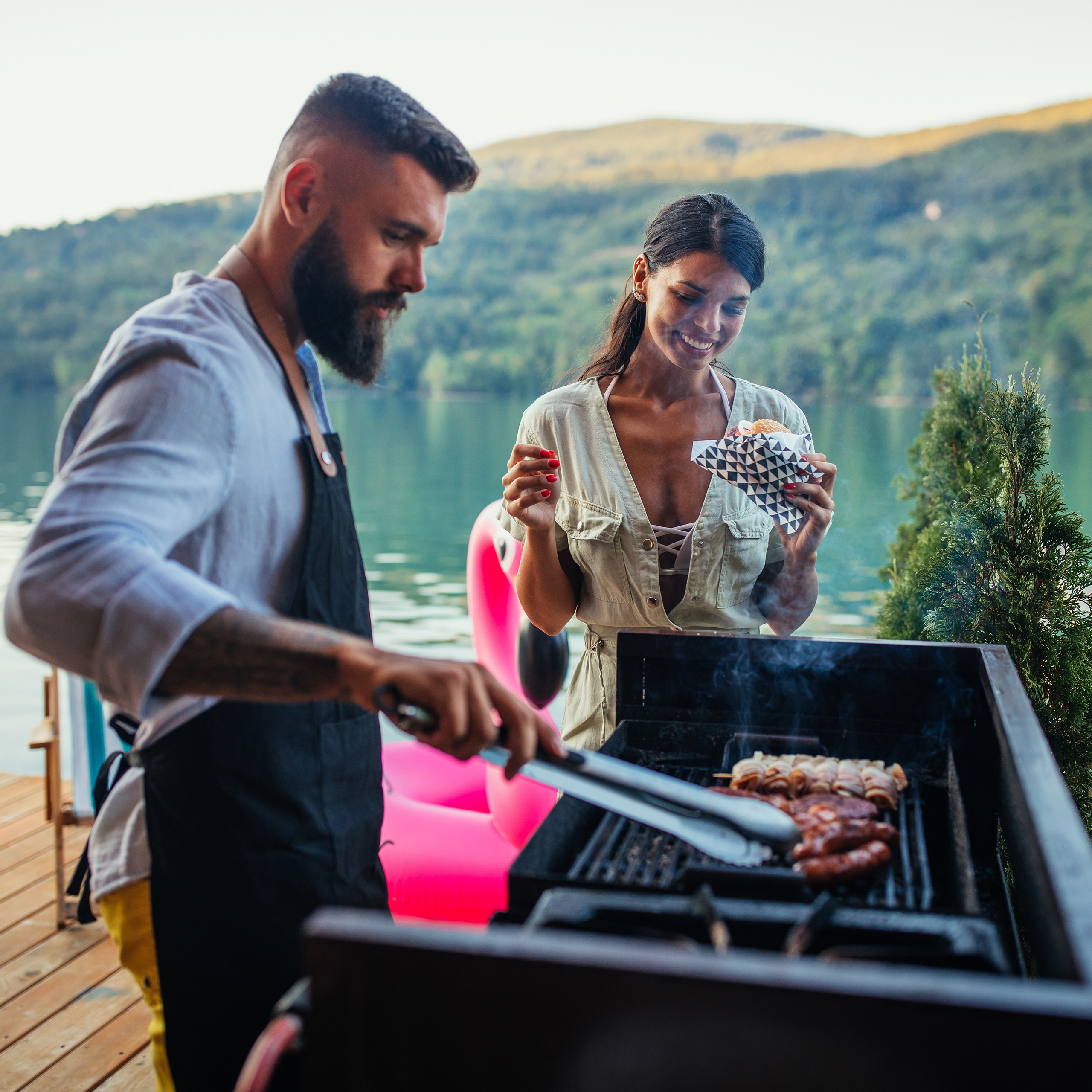 Ein bärtiger Mann grillt Fleisch, während eine Frau mit einem Burger daneben steht, an einem See.