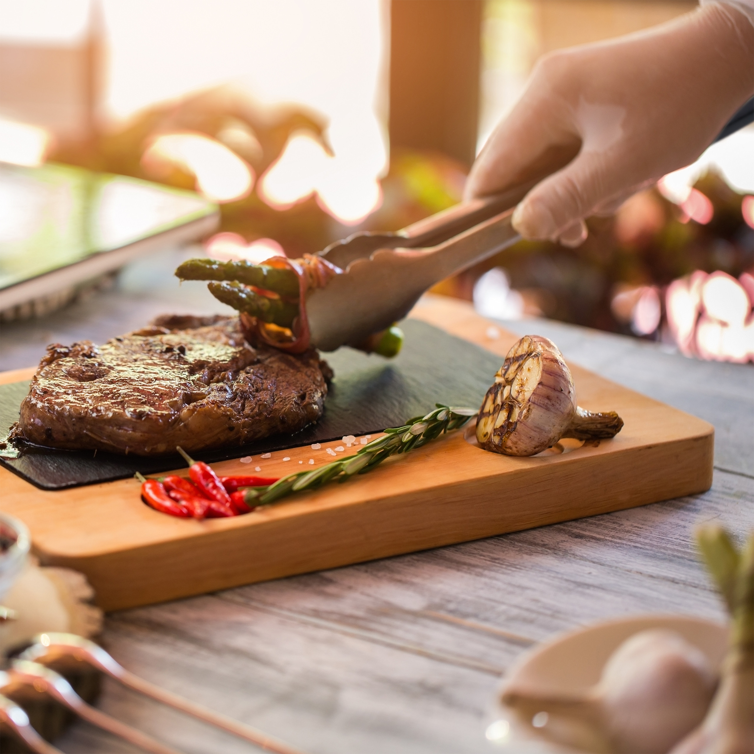 Ein Handschuh greift Spargel mit Zange neben Steak, Knoblauch und Chili auf Holzbrett.