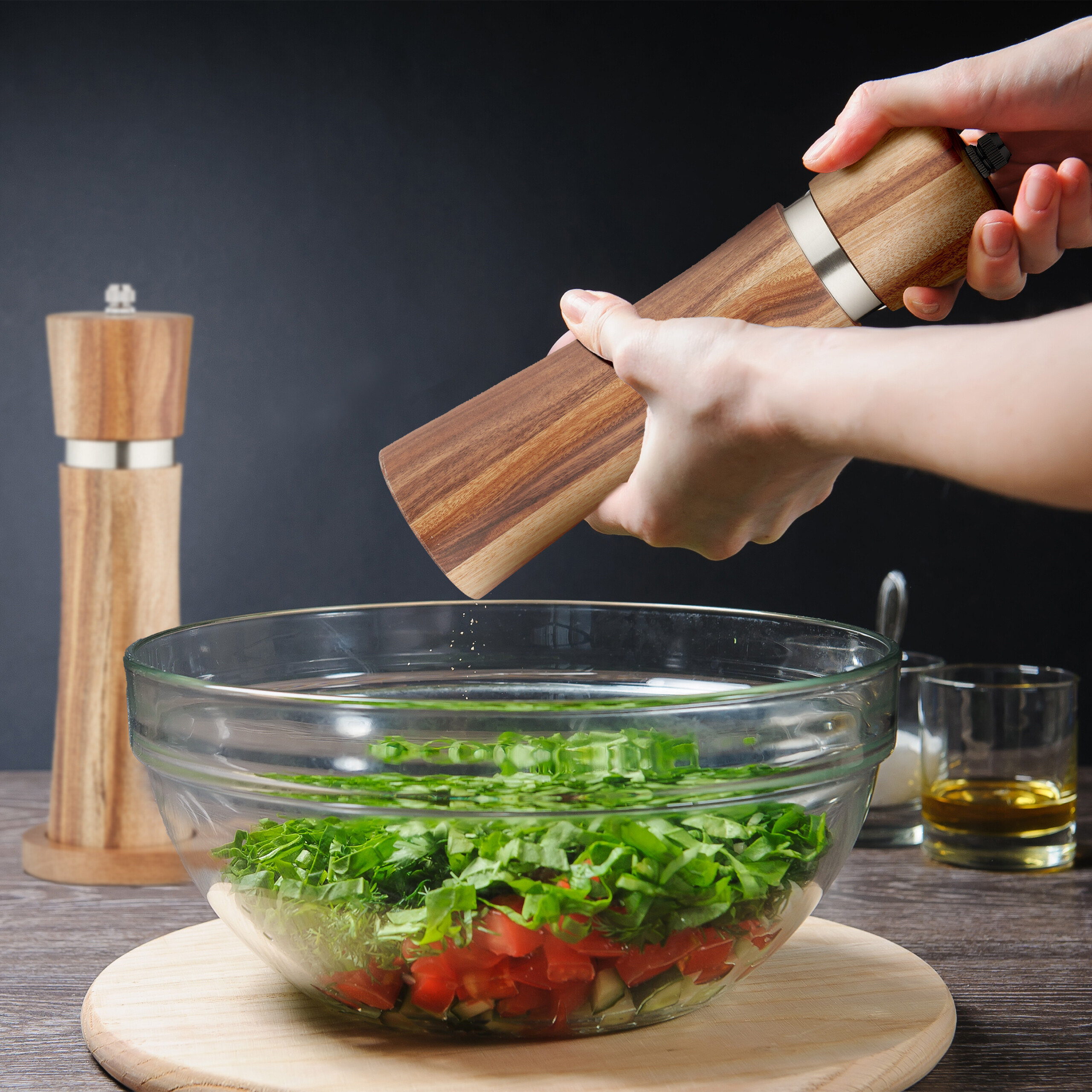 Person mahlt Gewürz aus einer Holzmühle über eine Glasschüssel mit Salat auf einem Holztisch vor dunklem Hintergrund.