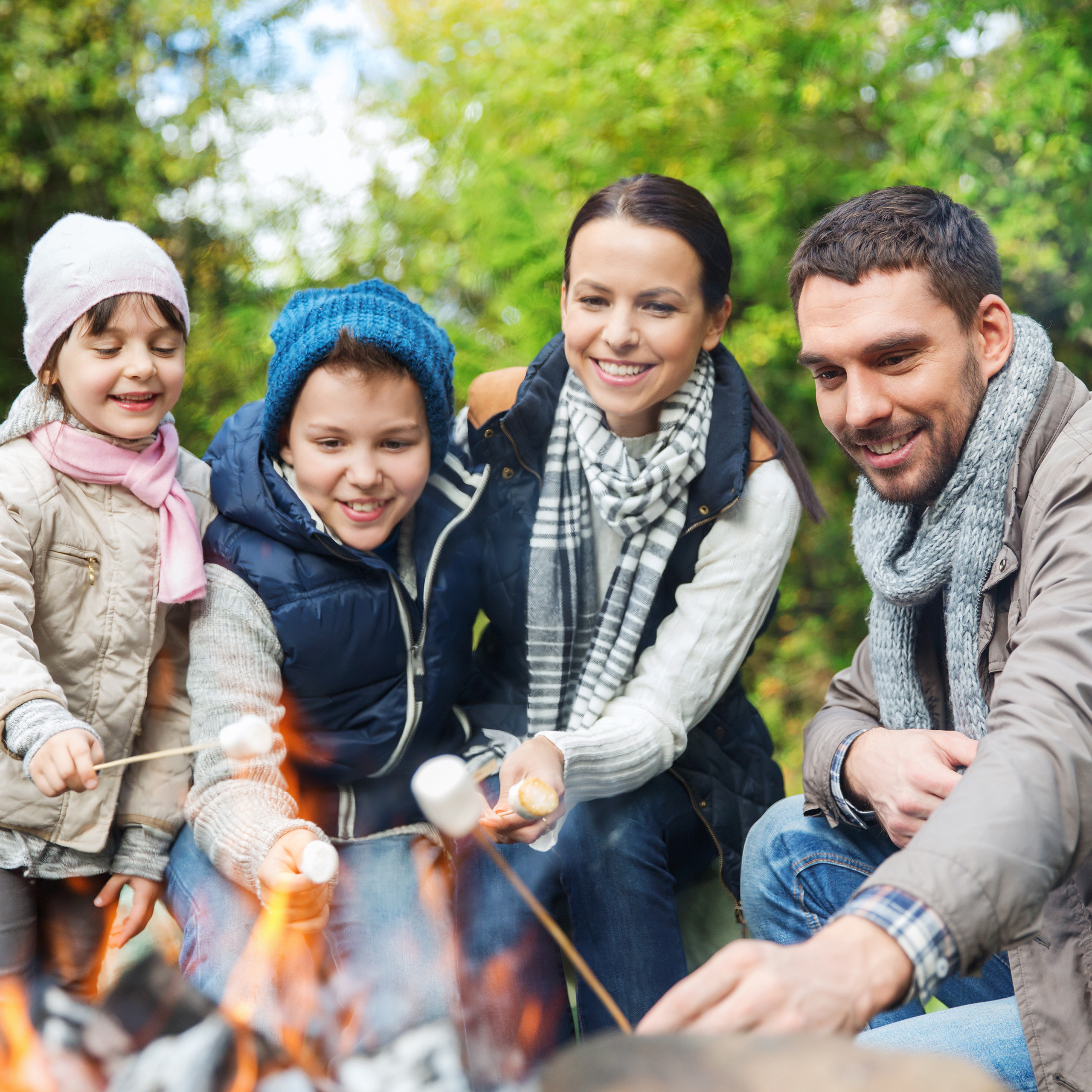 Eine lächelnde Familie mit zwei Kindern brät im Herbst draußen am Lagerfeuer Marshmallows.