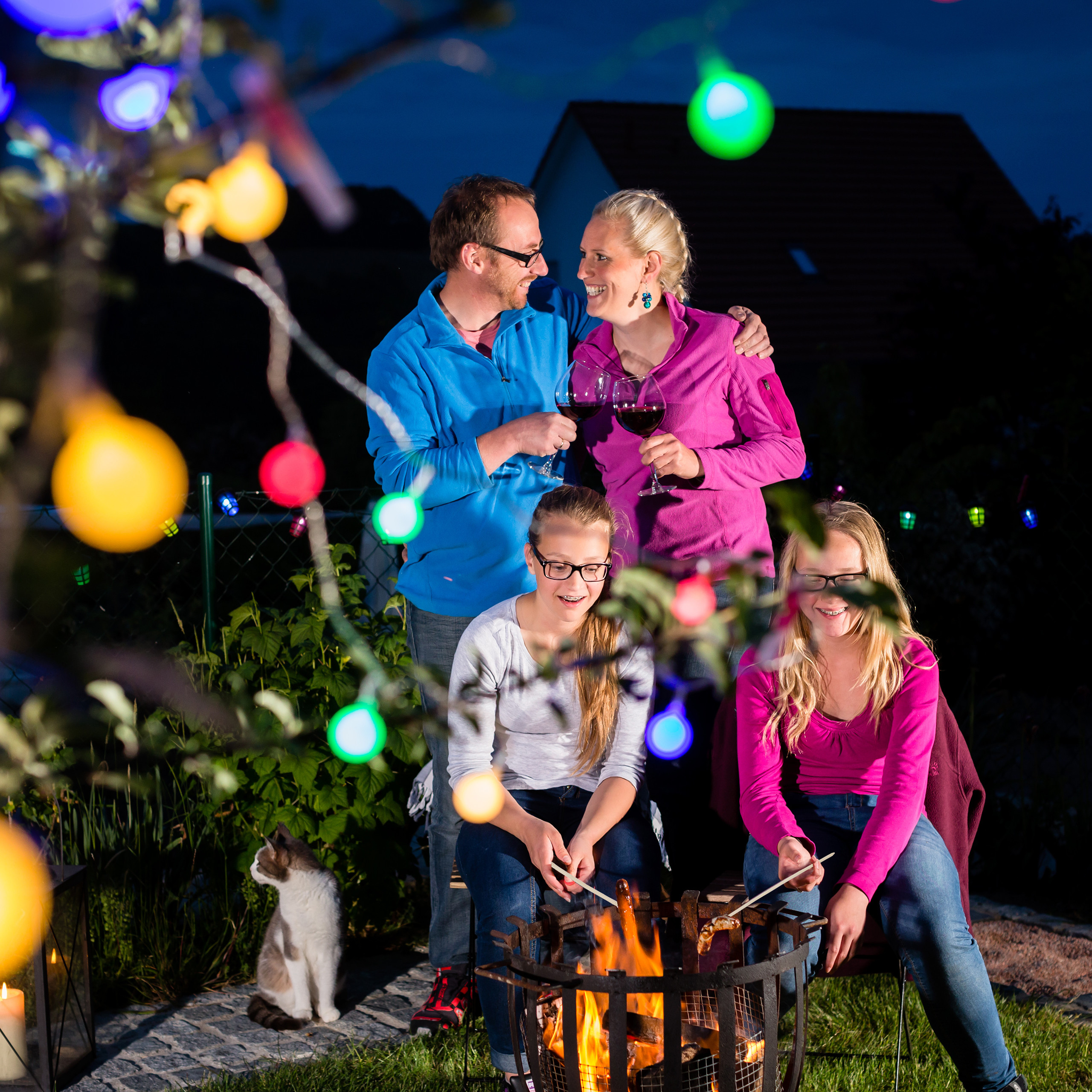 Eine Familie feiert nachts im Garten am Lagerfeuer mit Lichterketten und einem Katzenjungen.