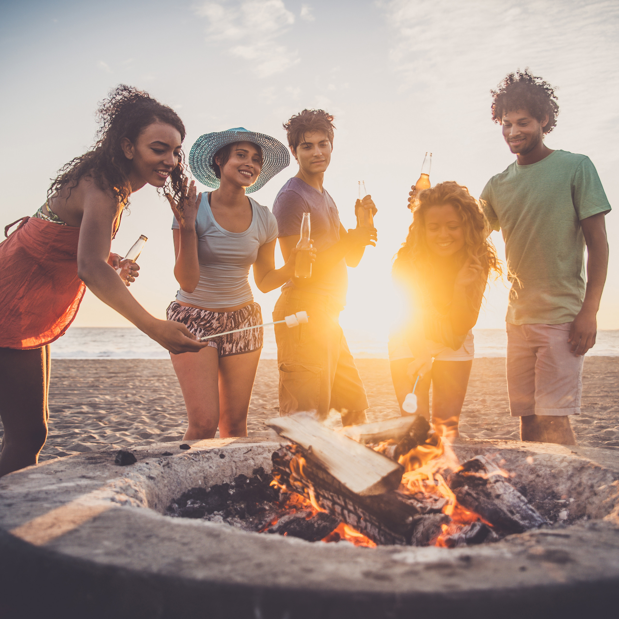 Fünf Freunde versammeln sich bei Sonnenuntergang um ein Lagerfeuer am Strand und rösten Marshmallows.