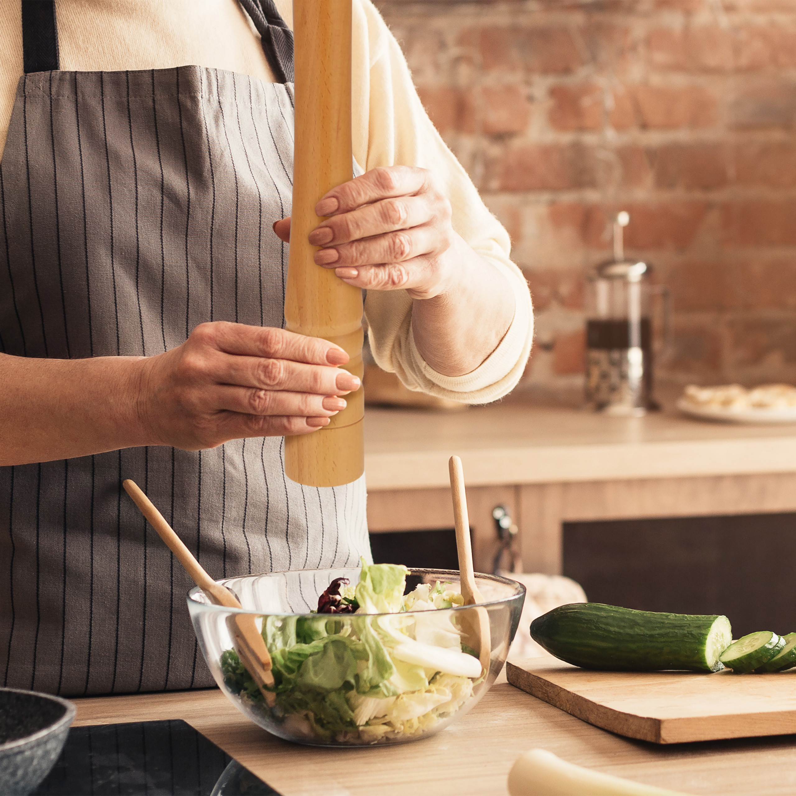 Eine Person in gestreifter Schürze würzt Salat in einer Glasschüssel mit einer Holzpfeffermühle.