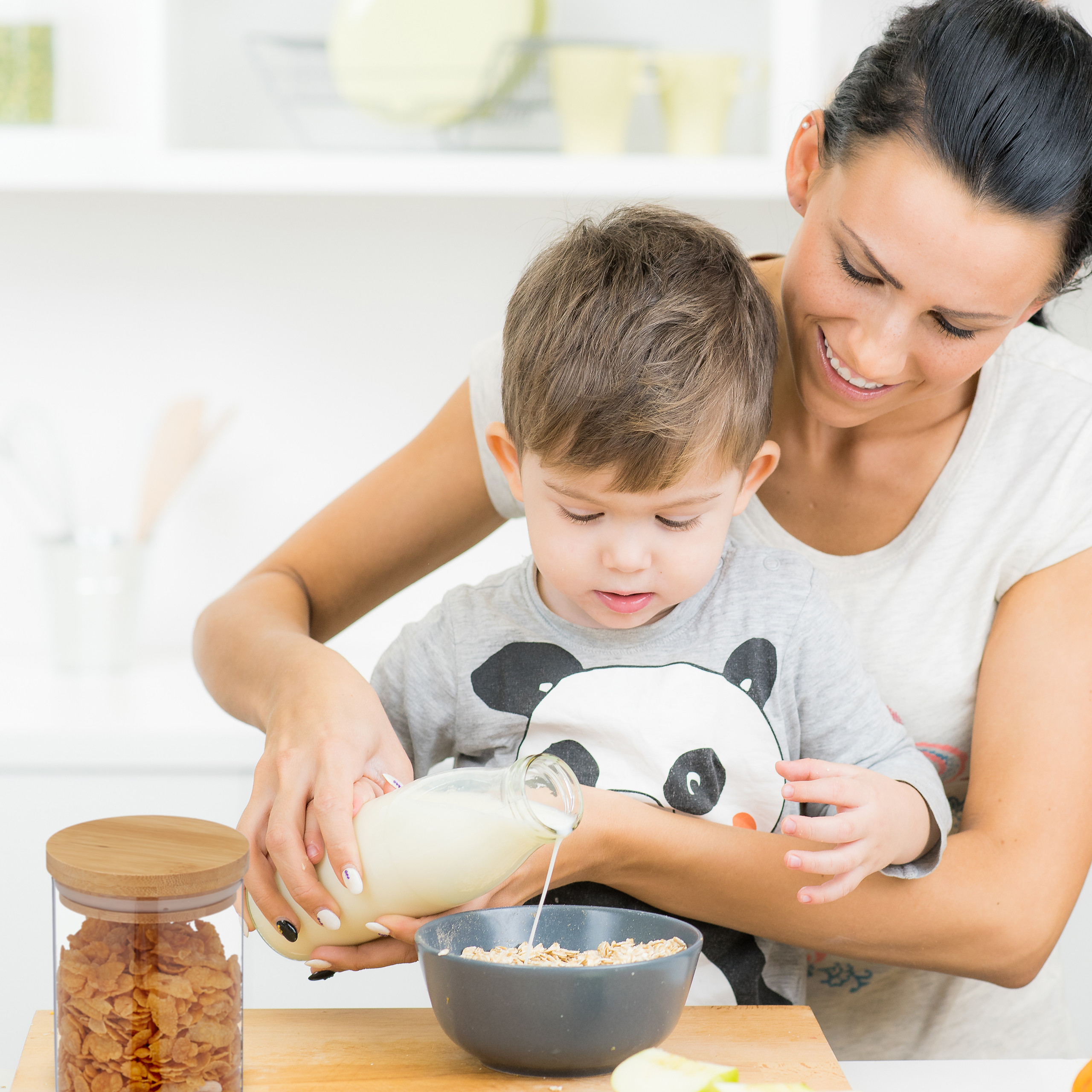 Eine Mutter und ein kleiner Junge gießen Milch in einer hellen Küche in eine Müslischüssel.