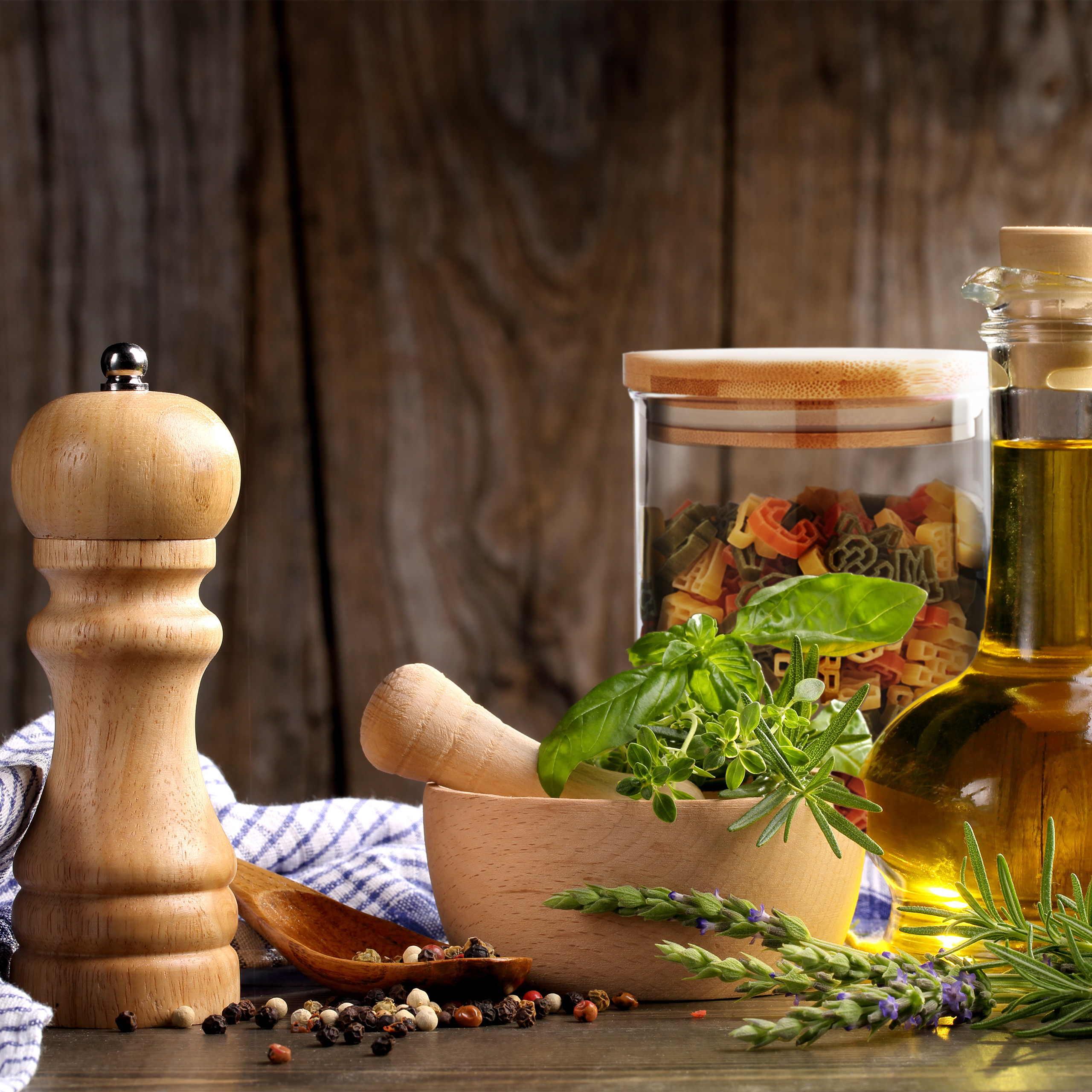 Holzmörser mit Kräutern, Pfeffermühle, Ölflasche und Nudeln auf Holztisch.