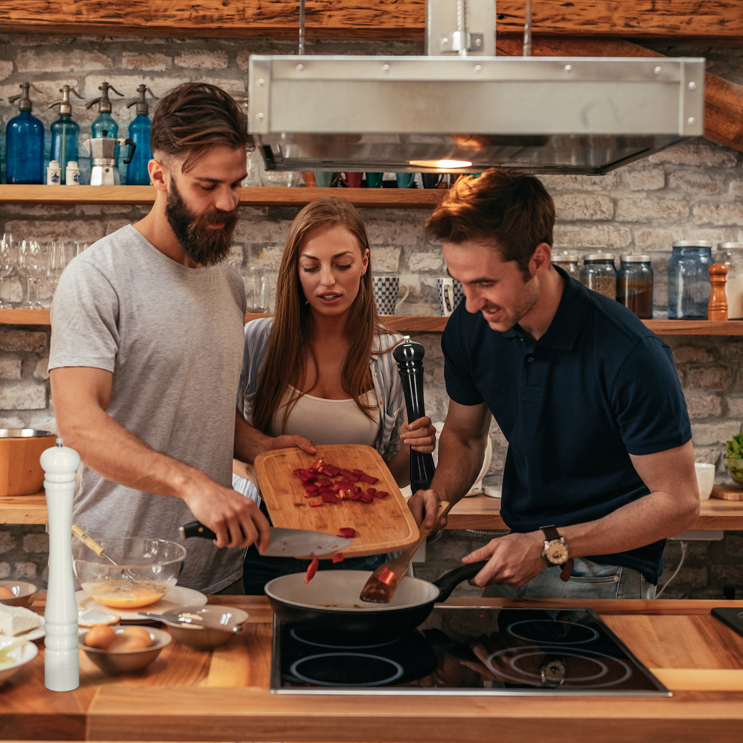 Drei Freunde kochen zusammen in einer rustikalen Küche und braten Gemüse an.