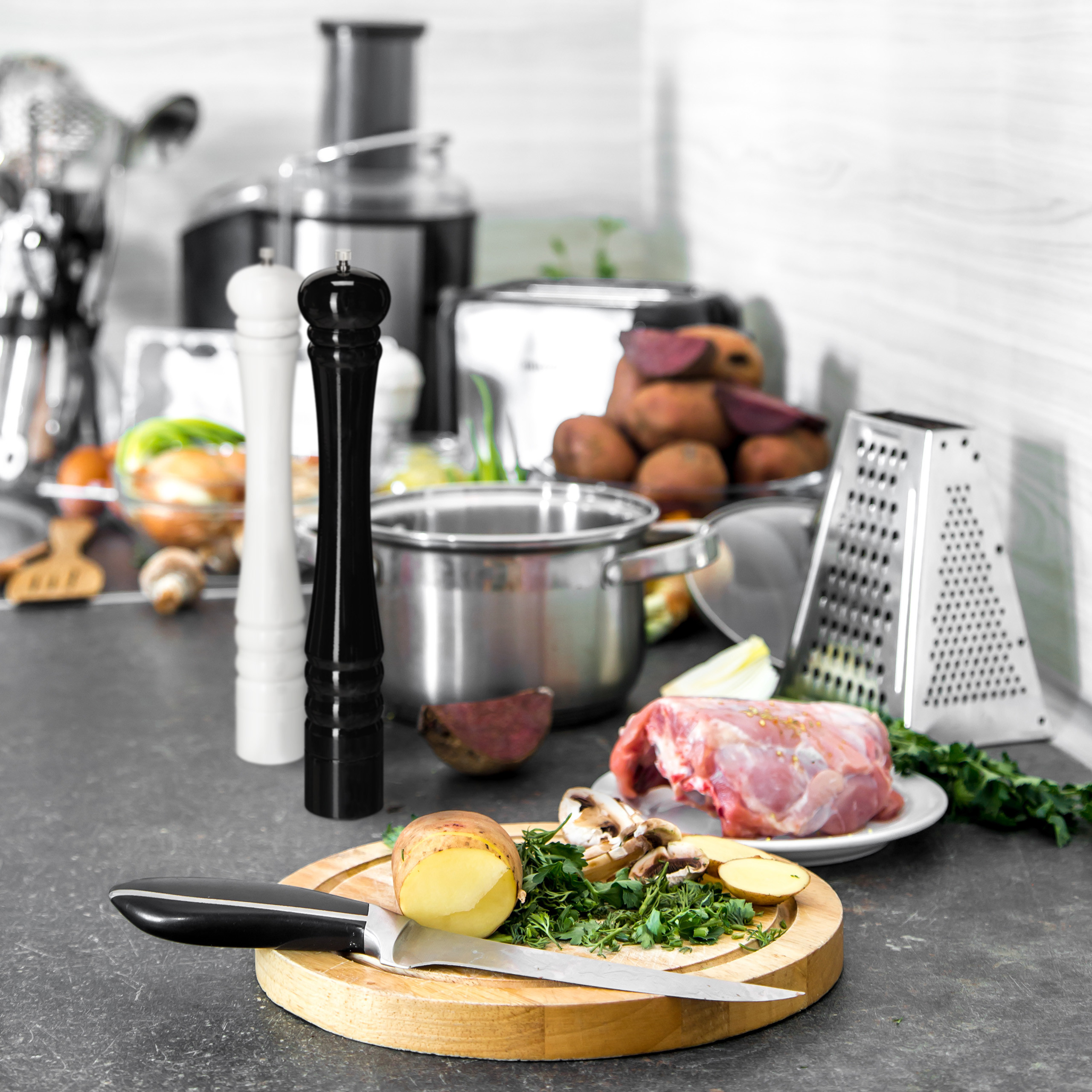 Küchenzeile mit Schneidebrett, Messer, Kartoffel, Kräutern, rohem Fleisch, Topf sowie Salz- und Pfeffermühle.