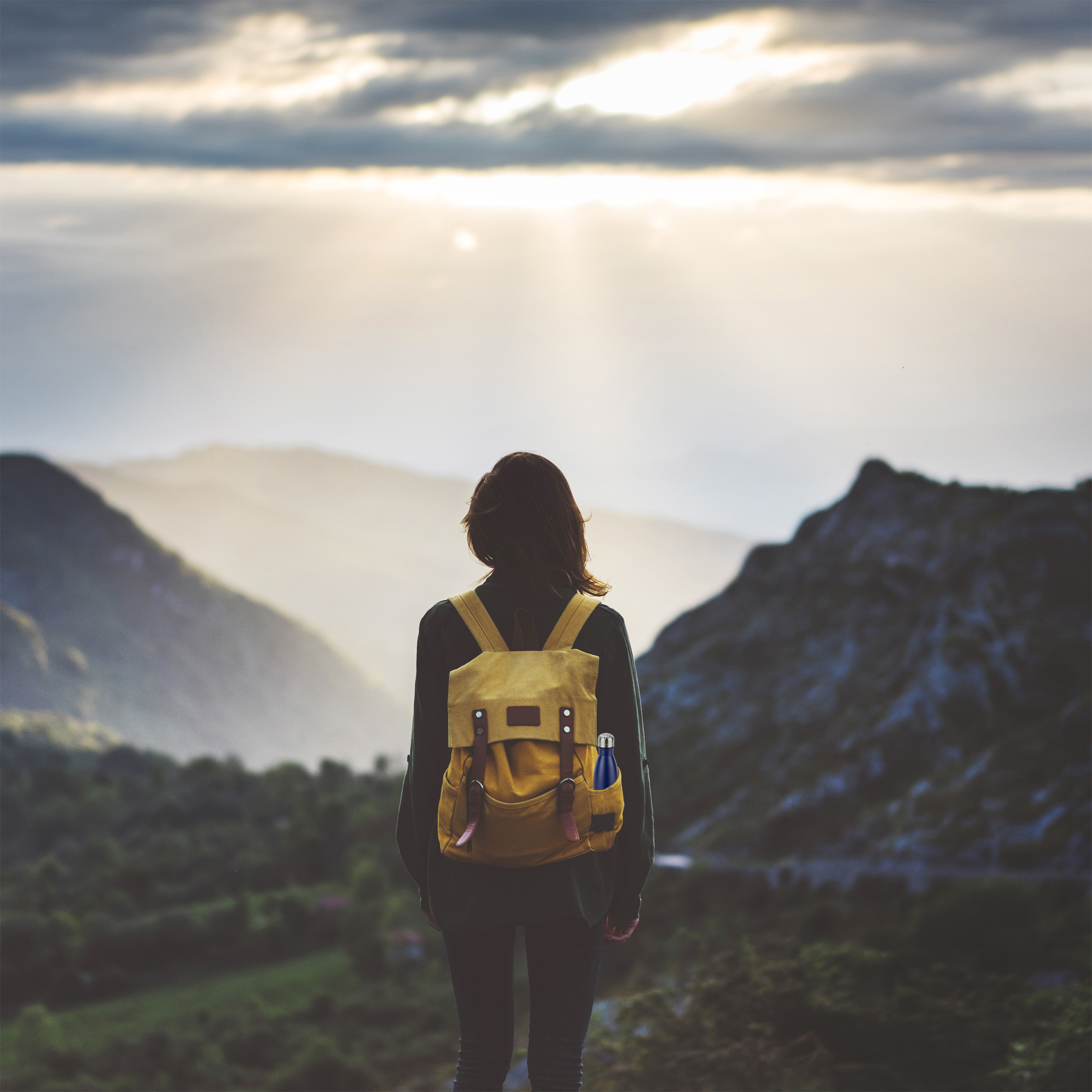 Eine Wanderin mit gelbem Rucksack blickt bei Sonnenuntergang auf eine Berglandschaft.