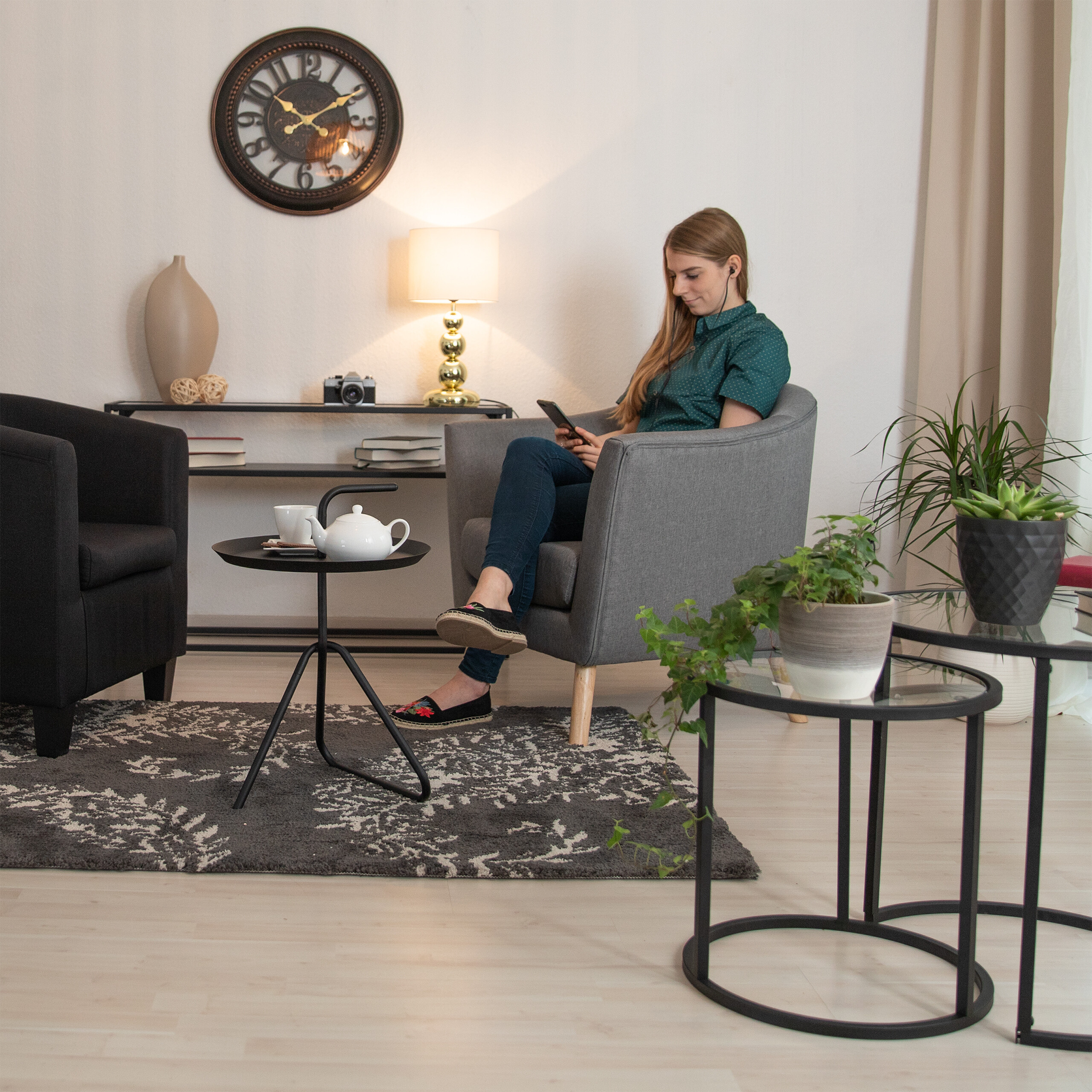Eine Frau sitzt in einem grauen Sessel in einem Wohnzimmer und nutzt ihr Smartphone, neben ihr ein Tisch mit Teekanne.