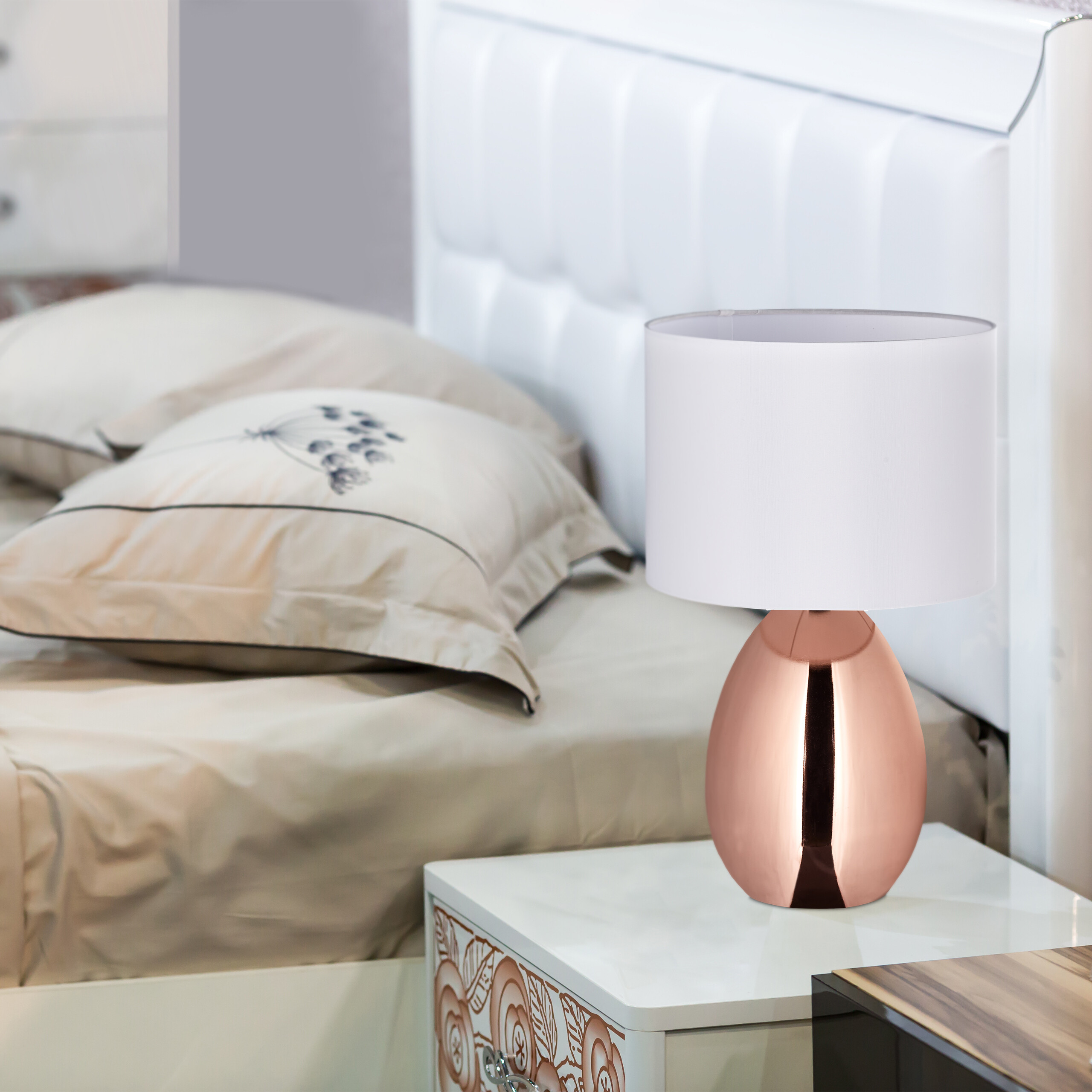Eine moderne Nachttischlampe in Roségold mit weißem Schirm steht neben einem gemachten Bett.