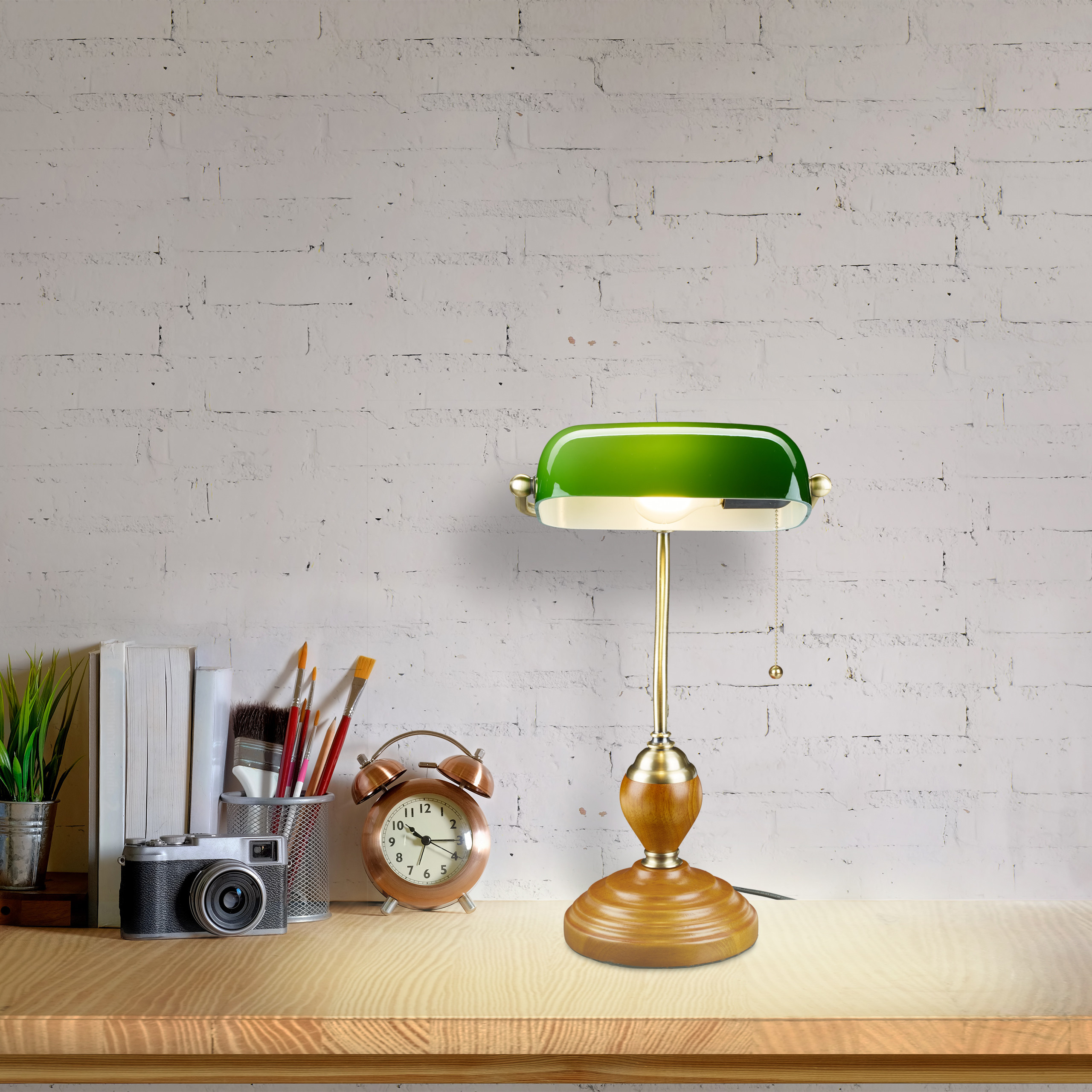 Eine grüne Bankerlampe steht auf einem Holztisch neben einer Kamera, Uhren und Büchern vor einer weißen Backsteinwand.