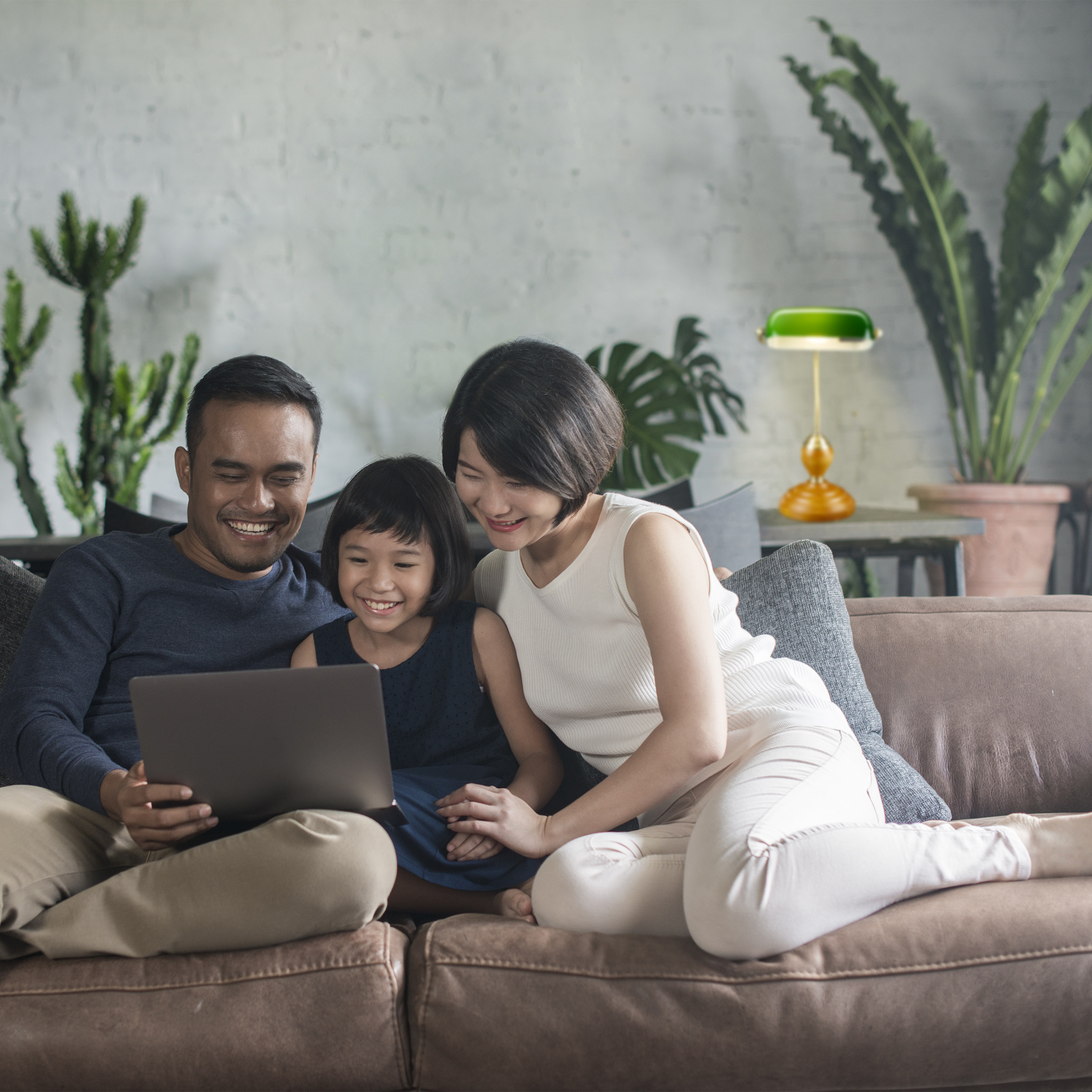 Eine asiatische Familie lächelt, während sie zusammen auf einem Sofa in ein Laptop schaut.