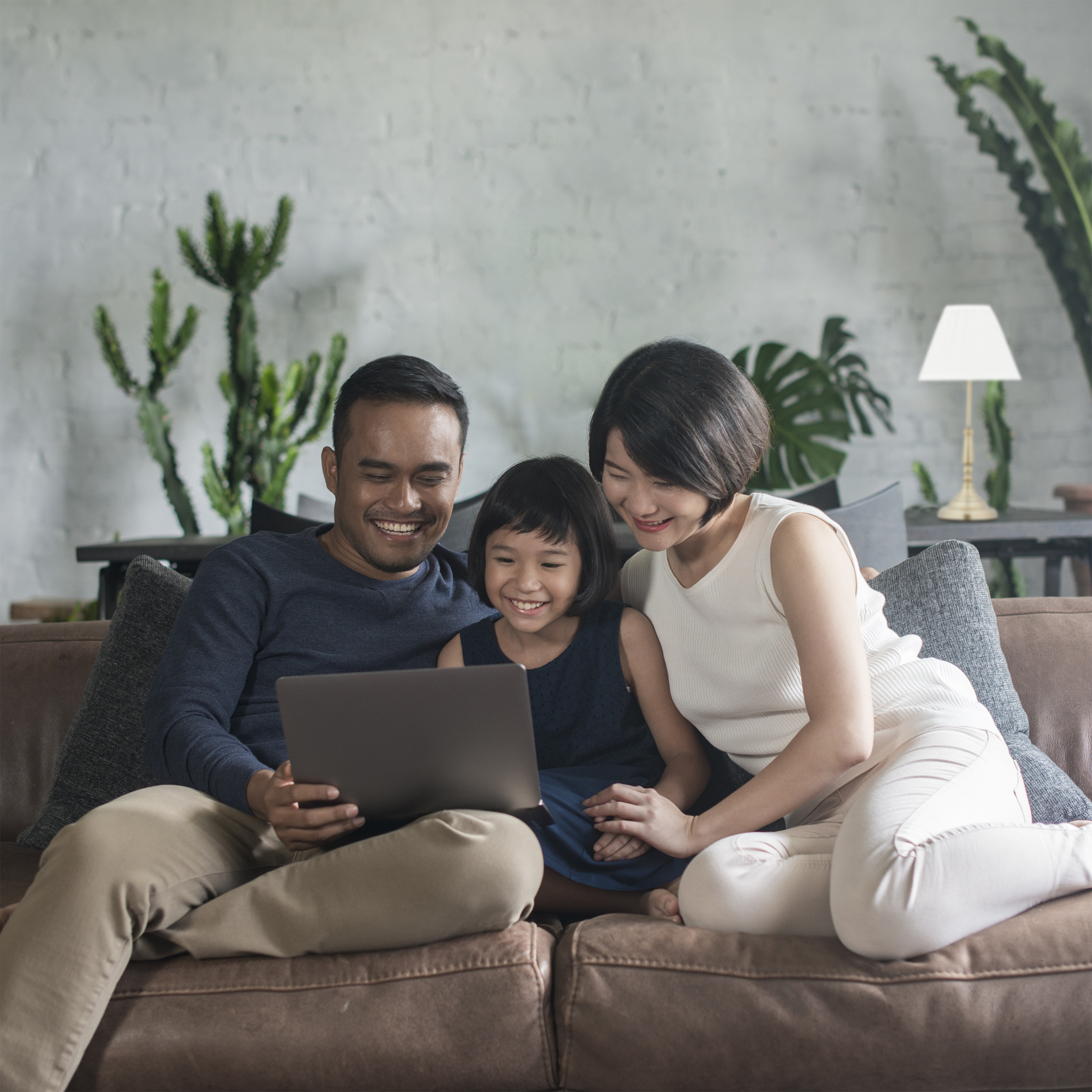 Eine lächelnde asiatische Familie sitzt auf einem braunen Sofa und schaut gemeinsam auf einen Laptop.