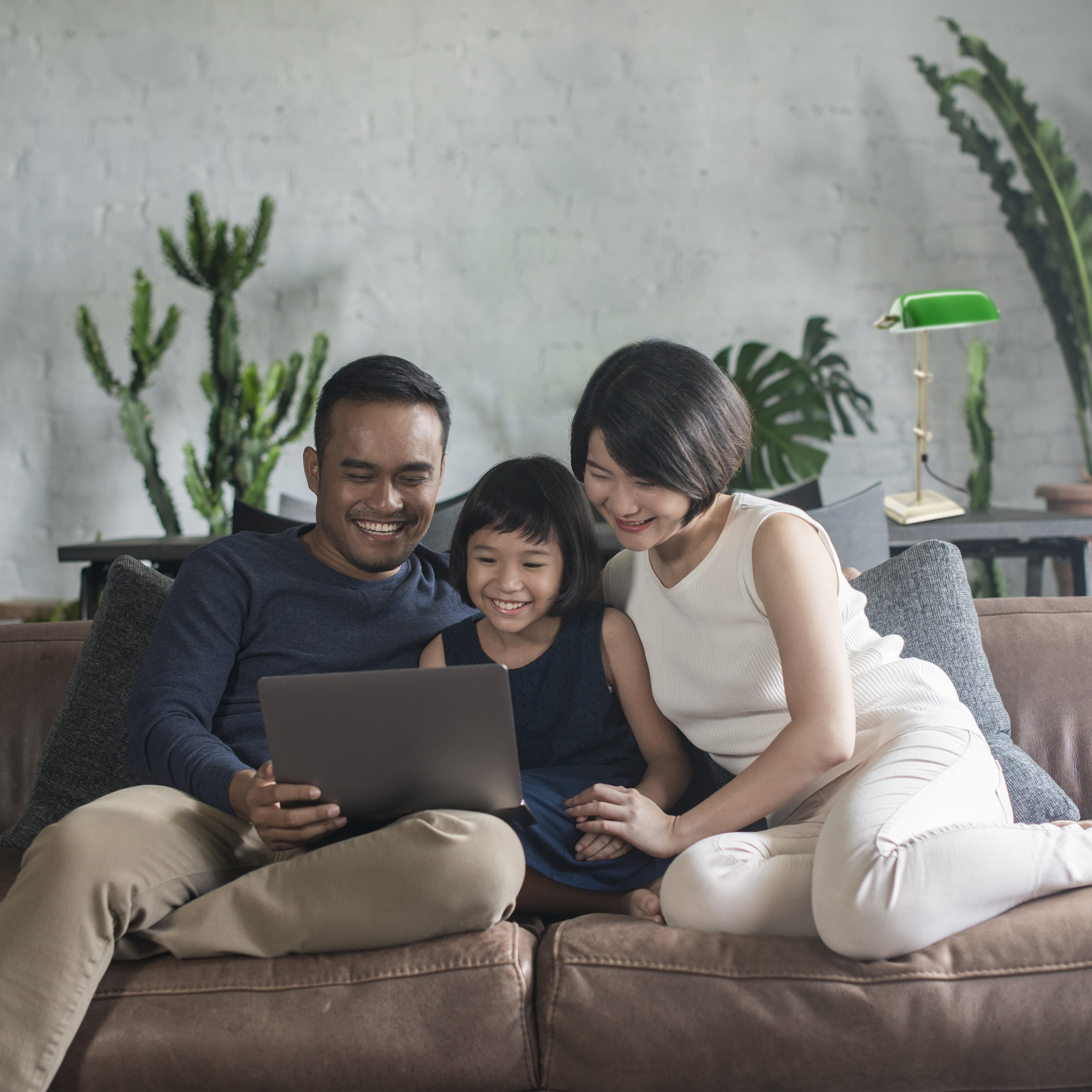 Eine lächelnde dreiköpfige Familie sitzt auf einem Sofa und schaut in einem hellen Raum auf einen Laptop.
